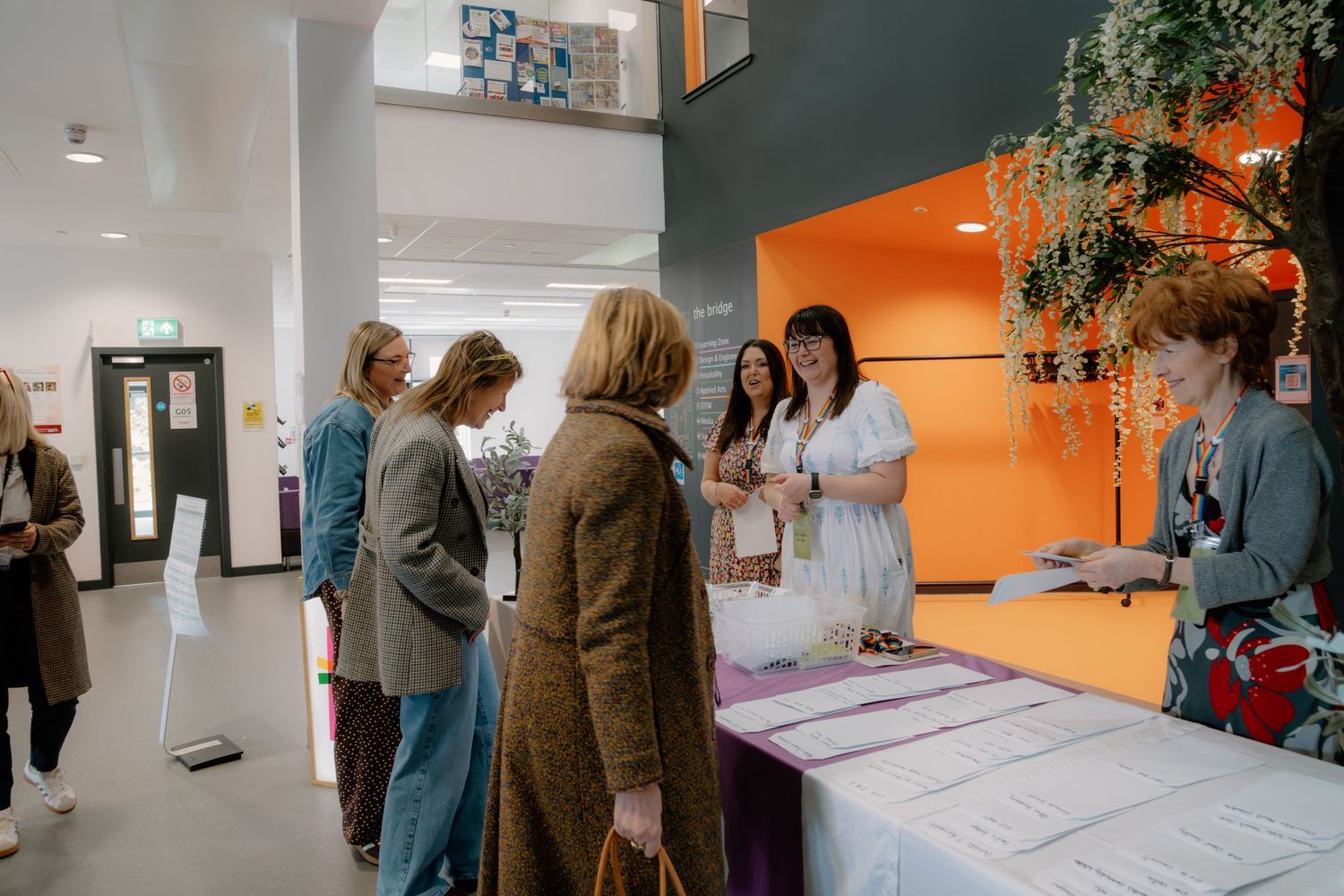 Women networking at a modern event registration desk with orange accent wall in a bright contemporary venue.