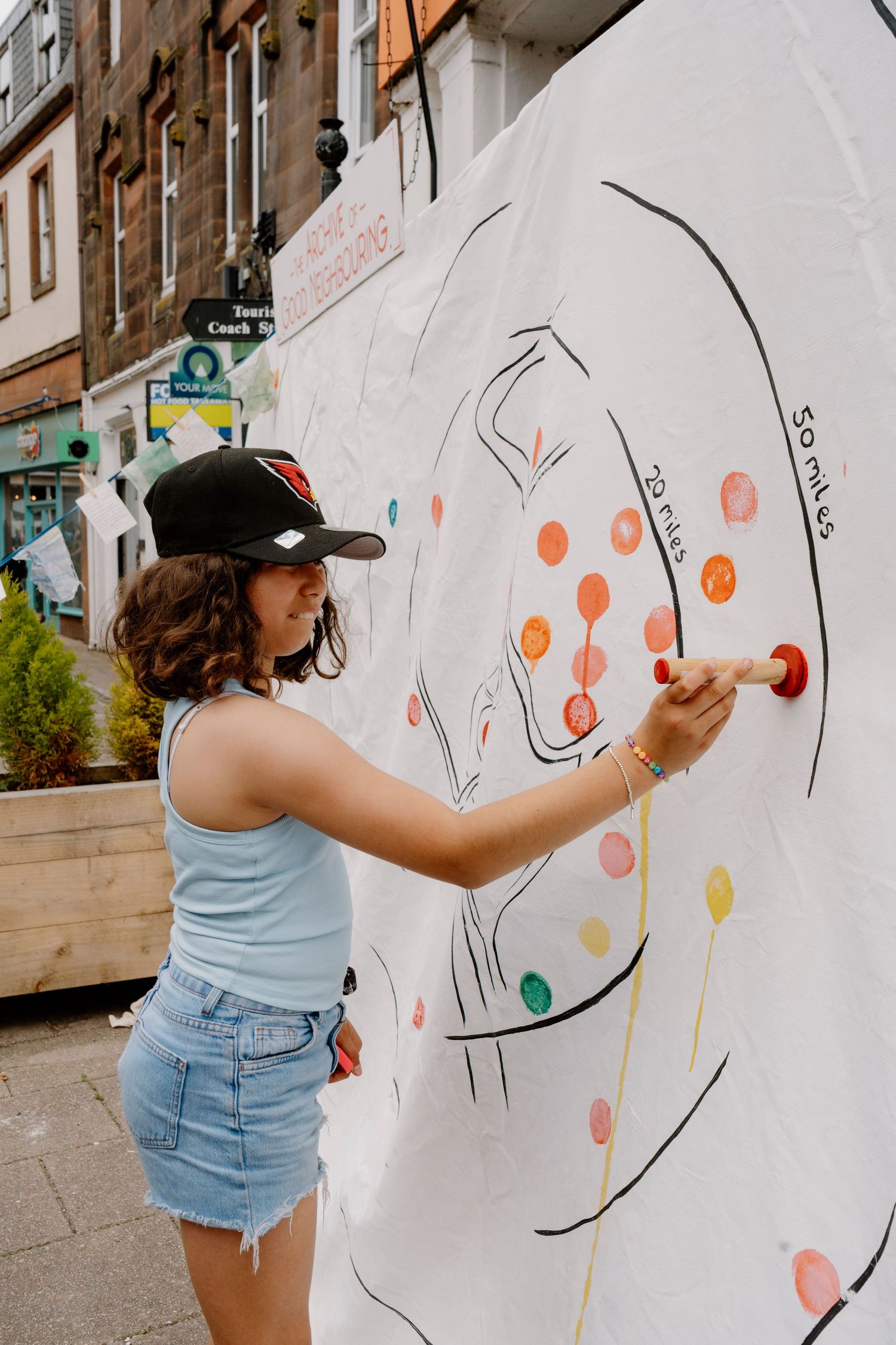 Young woman in cap and denim shorts adding colorful dots to a large outdoor mural on a city street.