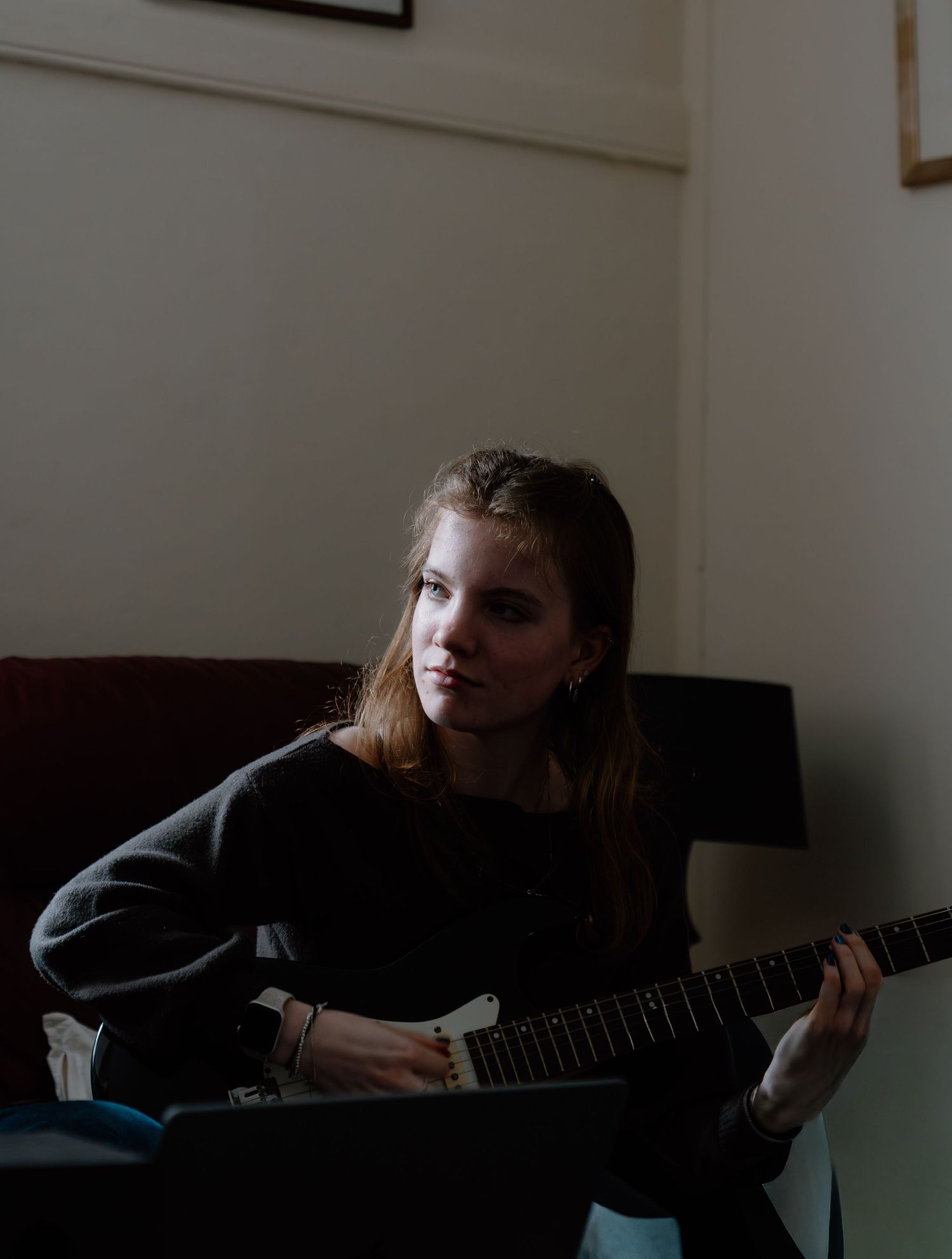 Young woman with long hair playing electric guitar while sitting in a dimly lit room.