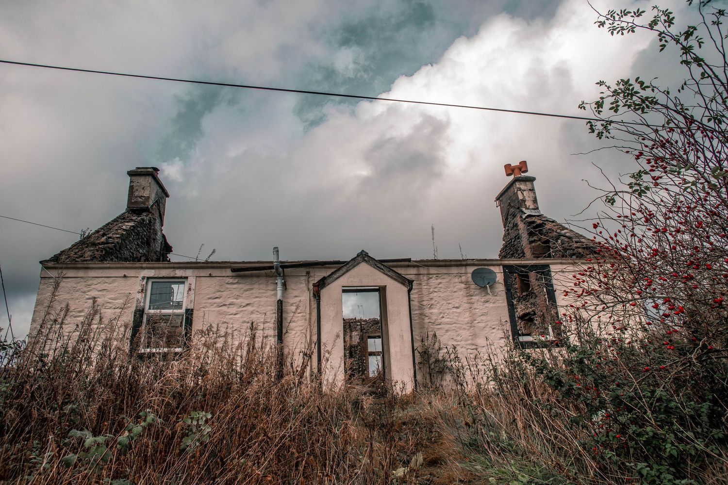 Panoramic view of an abandoned stone building with broken windows and crumbling chimneys against a cloudy sky.
