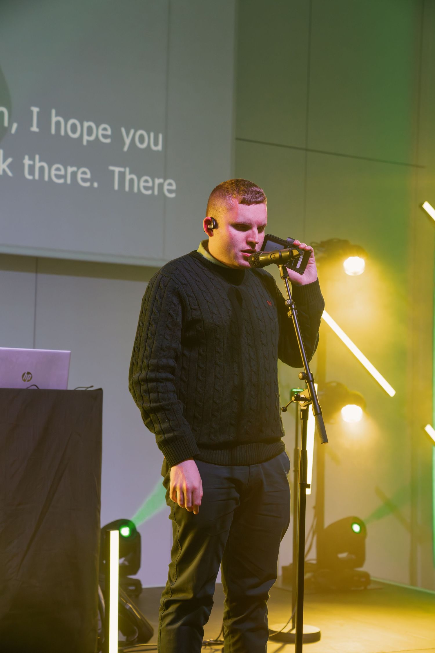 Young man in black sweater singing into microphone on stage with green lights and lyrics projected on screen behind him.