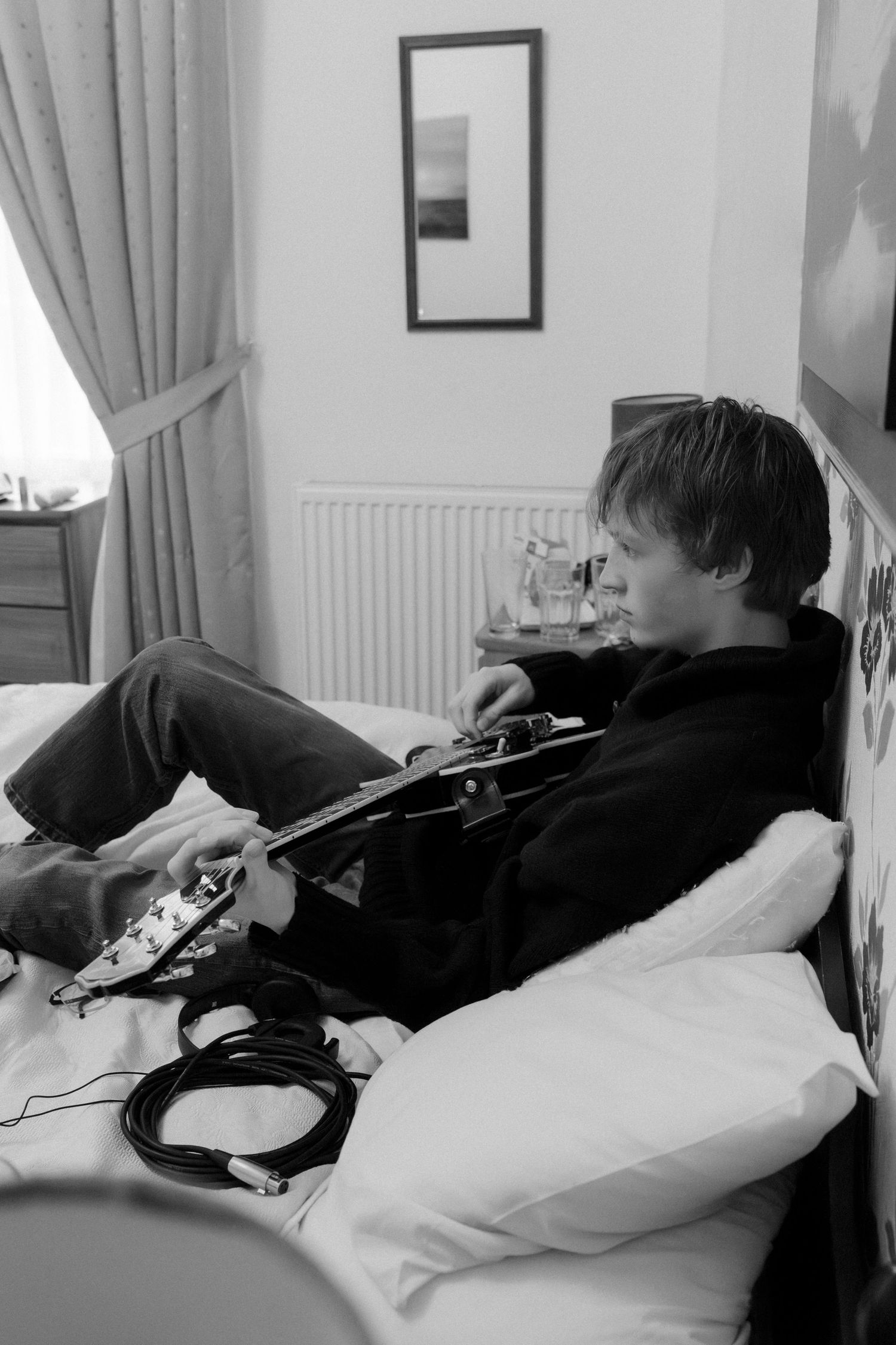 Young man playing electric guitar on bed in black and white photo with headphones and cables nearby.