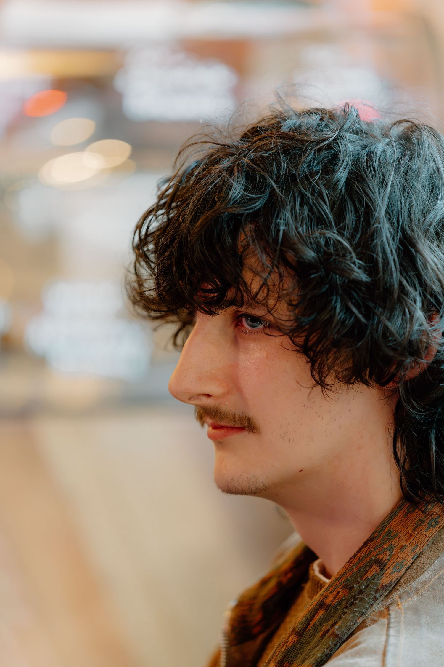 Young man with curly dark hair and mustache photographed in profile against a blurred bokeh background.