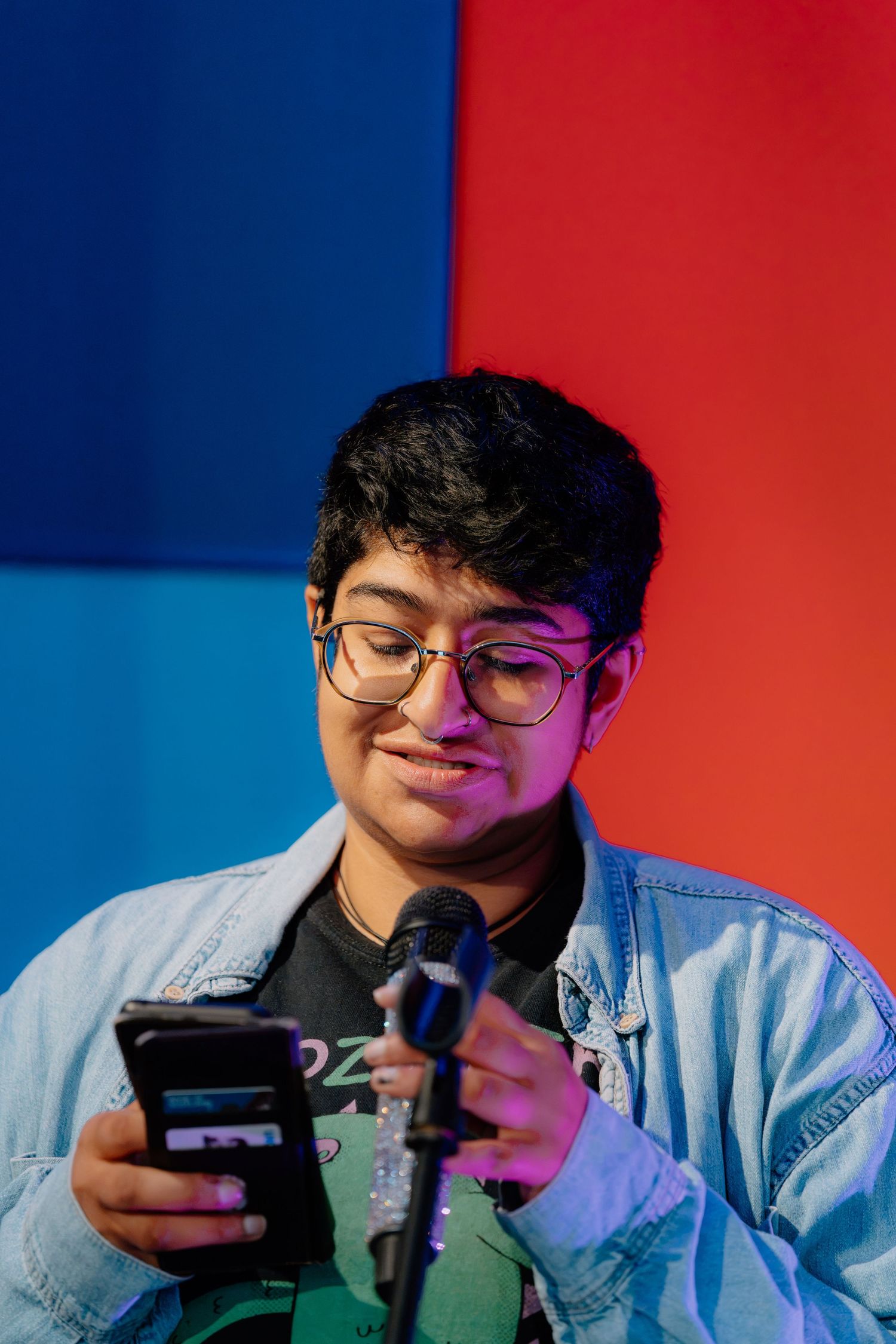 Young person with glasses smiling at phone near microphone, blue and red background.
