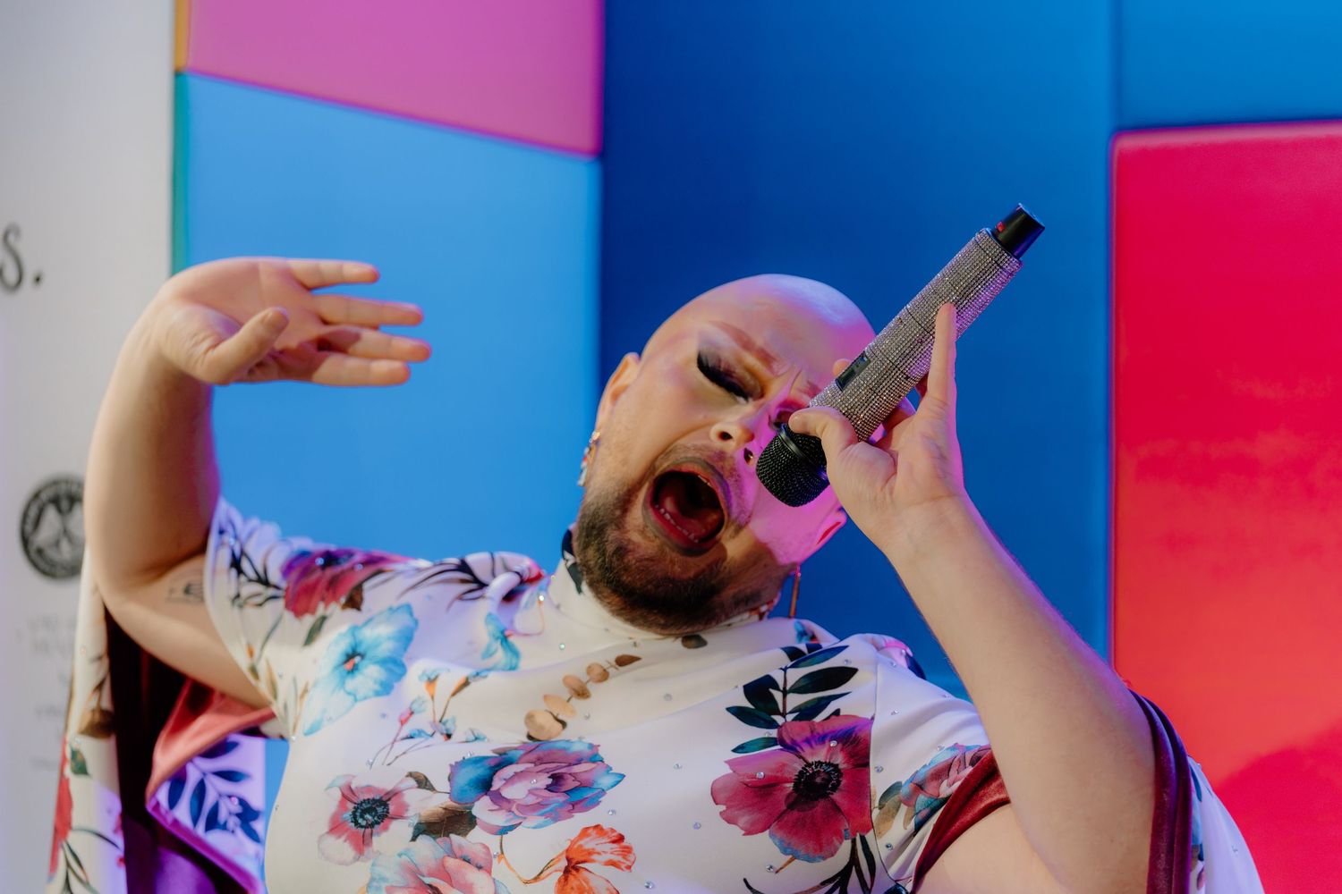 Bald performer in floral shirt singing passionately into microphone on colorful stage.