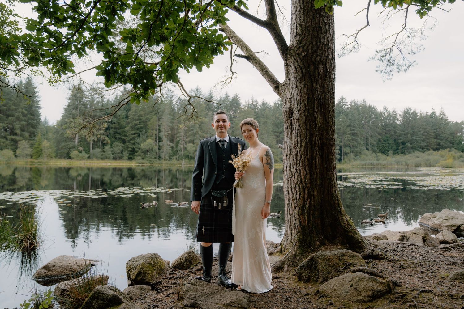 Bride and groom posing by a lakeside tree, groom wearing a Scottish kilt, bride in elegant gown, lush forest backdrop.