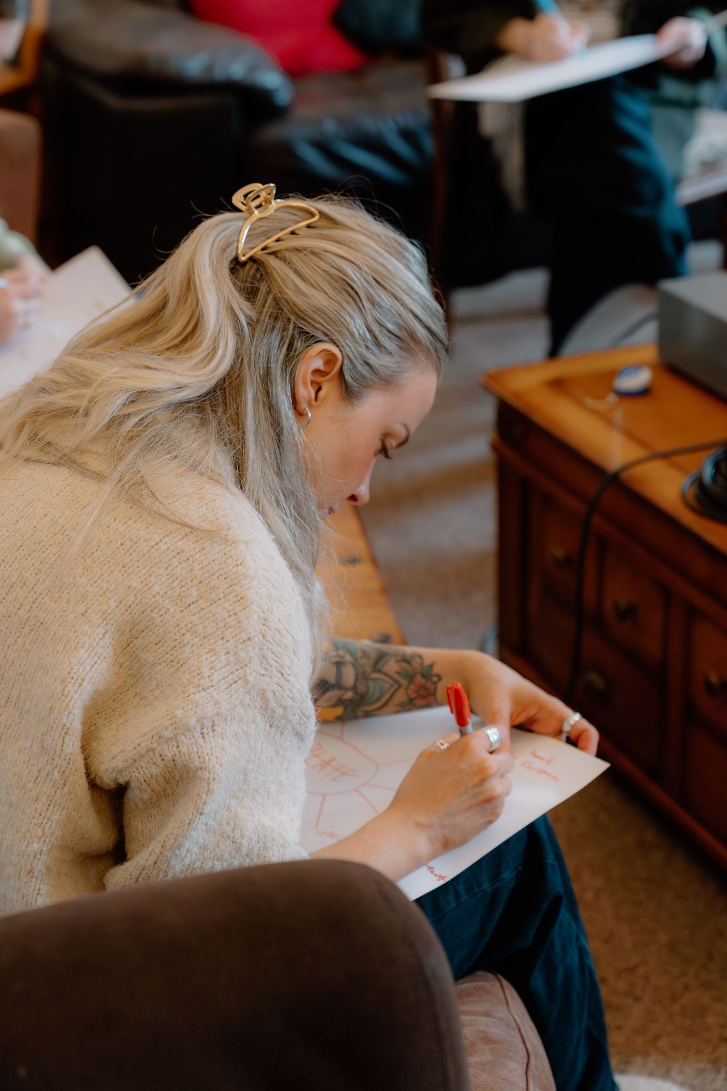 Blonde woman with tattoos and hair clip writes with red marker on paper in a casual group setting.