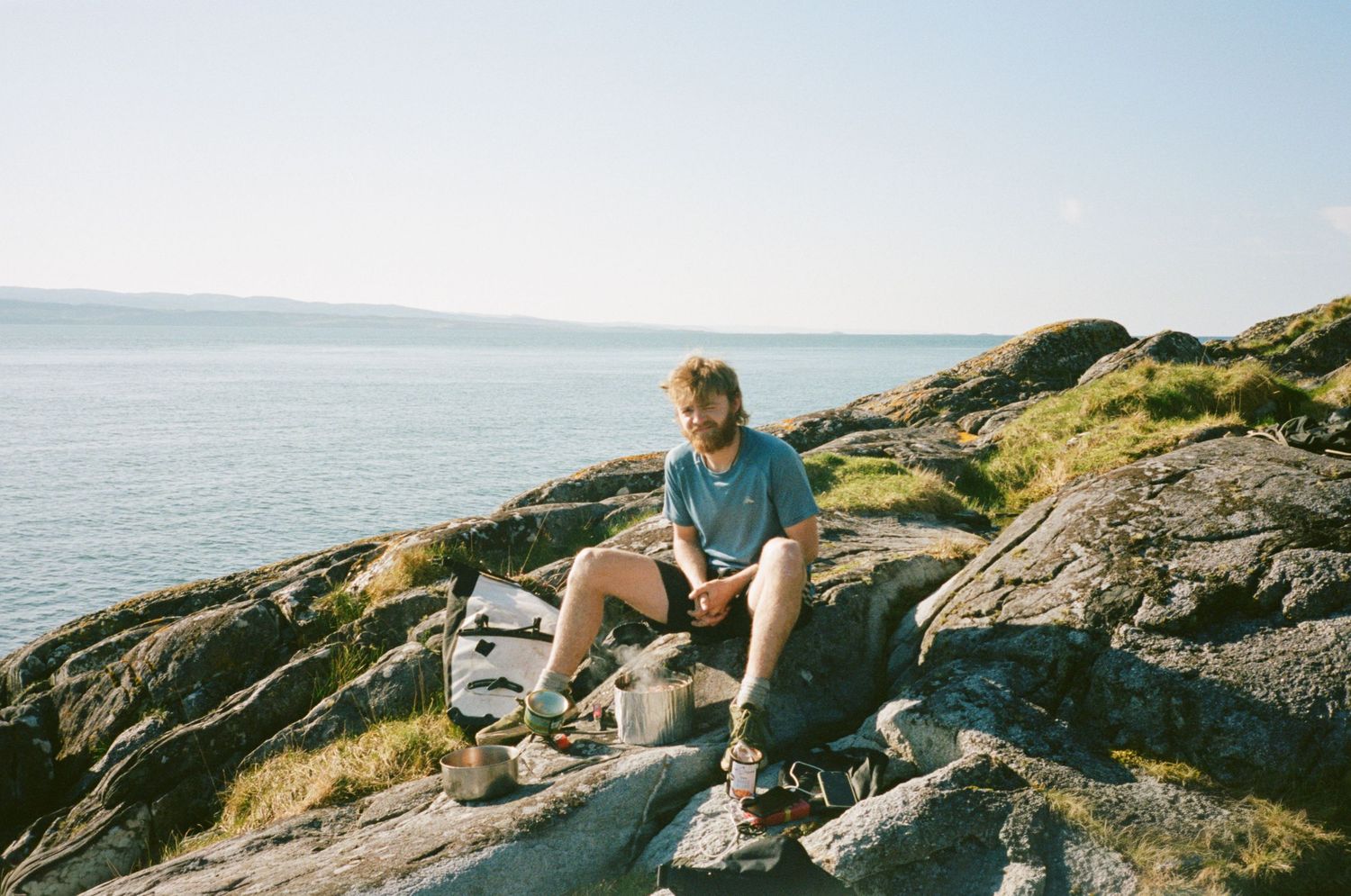 Man sitting on rocky coastal cliffside wearing blue shirt and shorts, with calm sea and clear sky behind him.