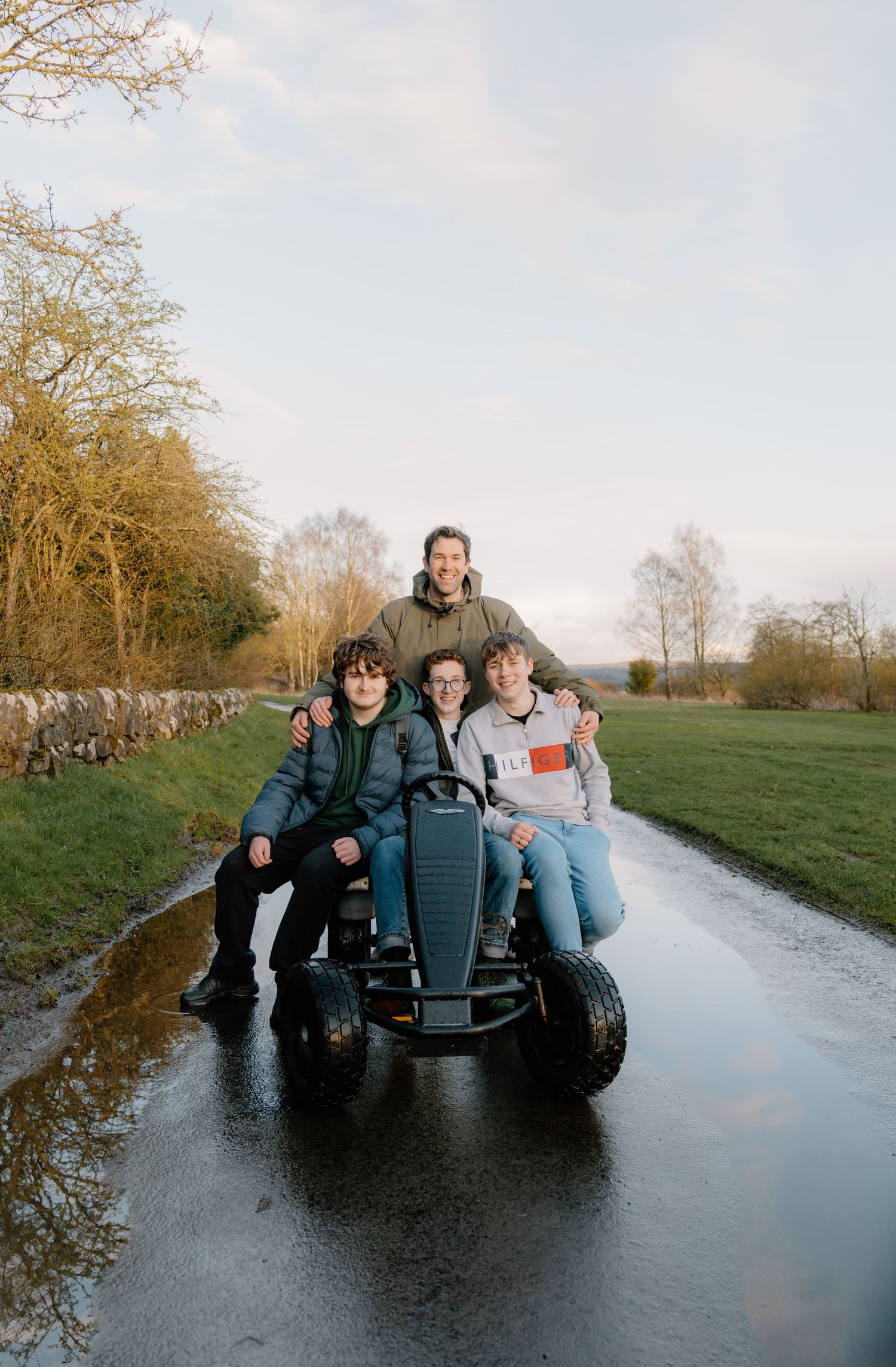 Four people smiling on a go-kart on a wet rural path surrounded by green fields and trees.