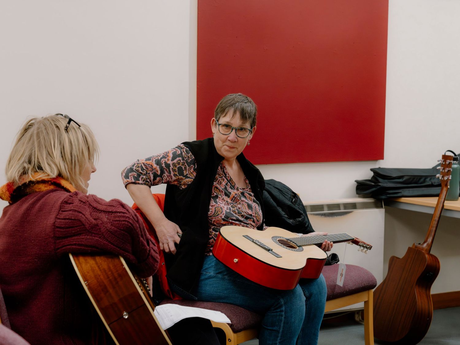 Two women sitting with acoustic guitars in a music room with a red painting on the wall.