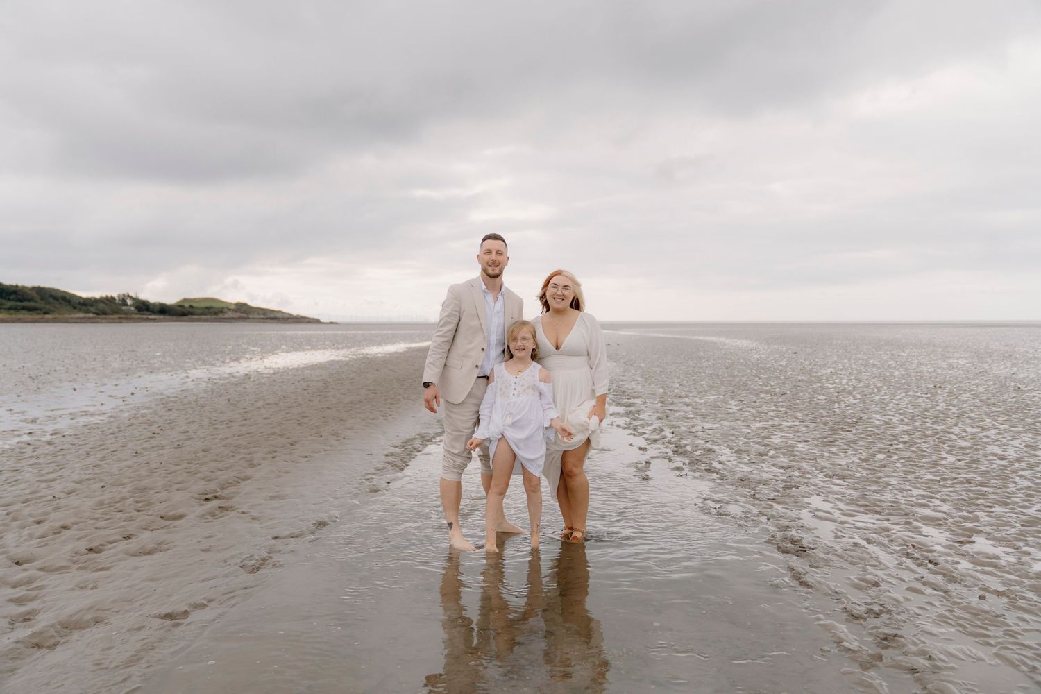 A couple in light colored clothing walks along a misty beach shoreline at sunset.