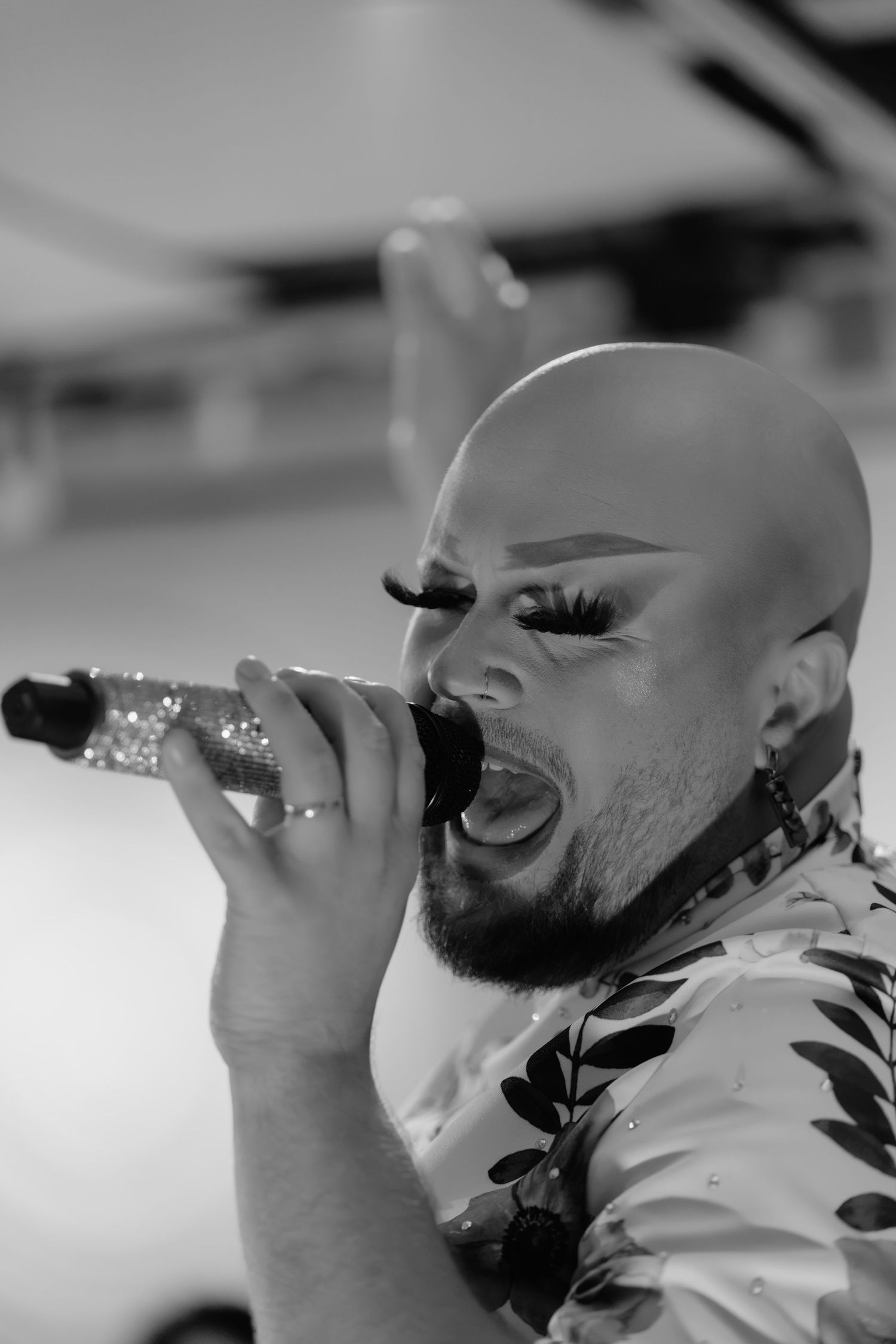 Bald drag performer with dramatic eye makeup and beard singing passionately into a glittery microphone, black and white photo.