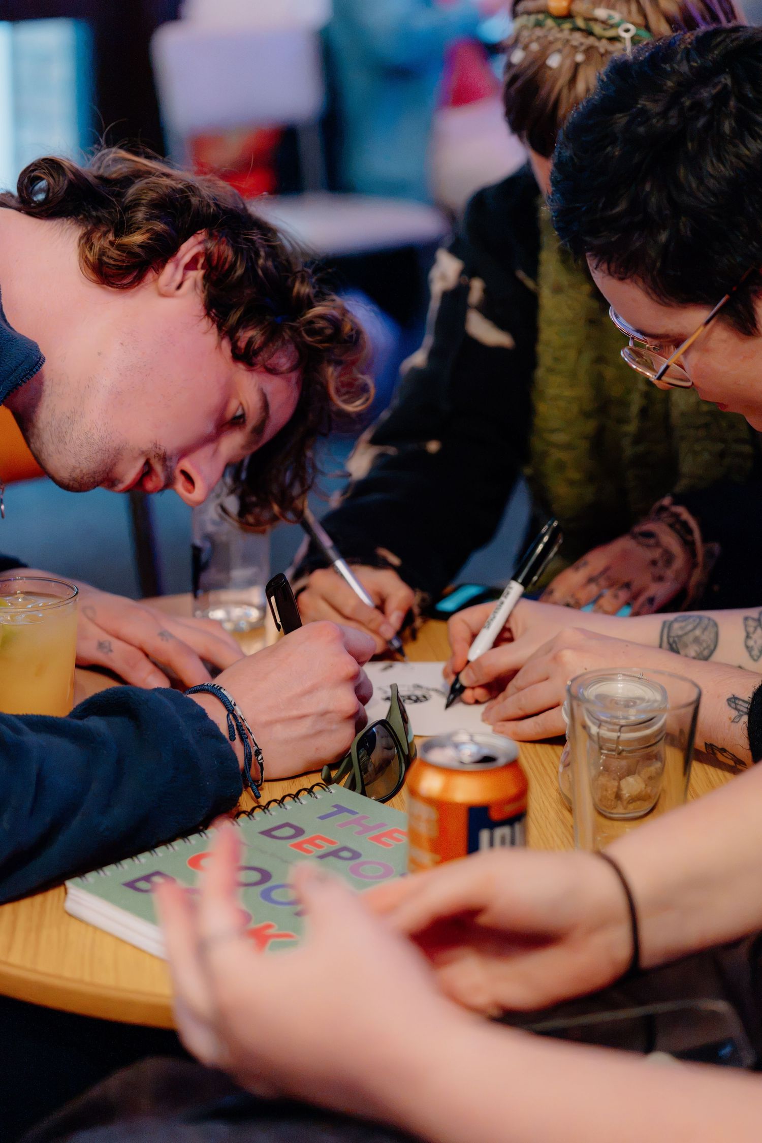 Group of friends leaning over a table, writing on a notepad with drinks nearby in a lively bar setting.