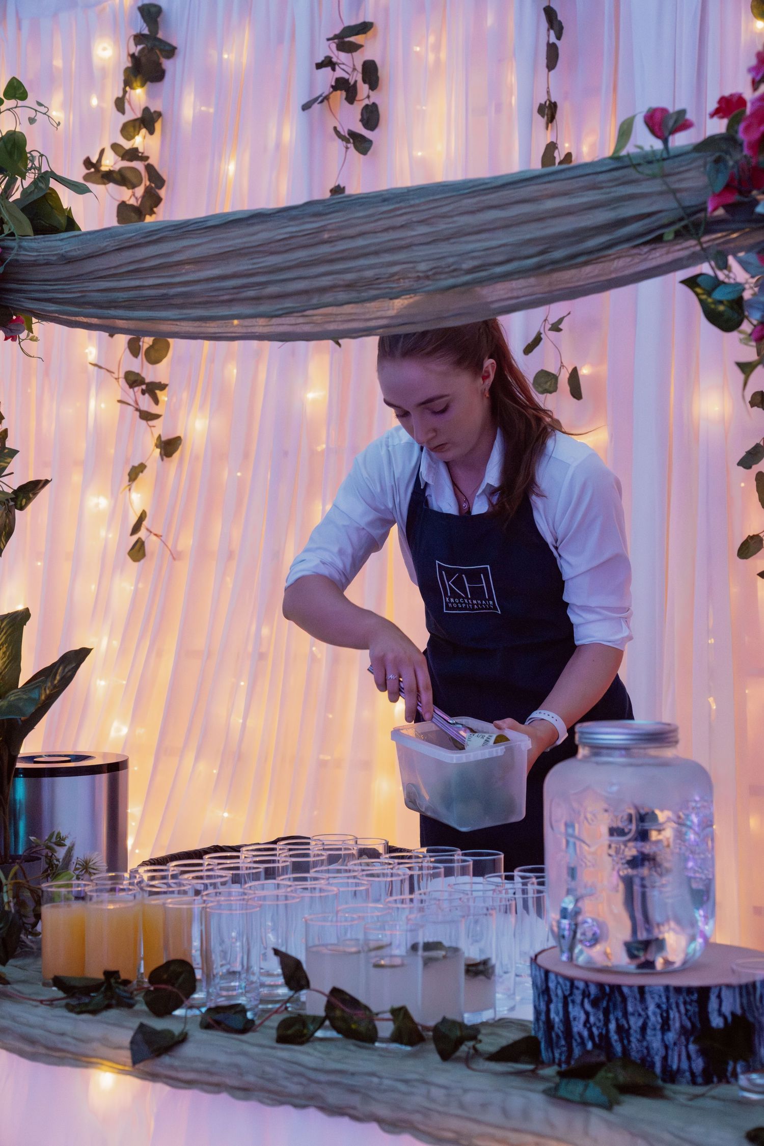 Bartender in black apron pouring drinks at elegant event bar with fairy lights and glass dispensers.