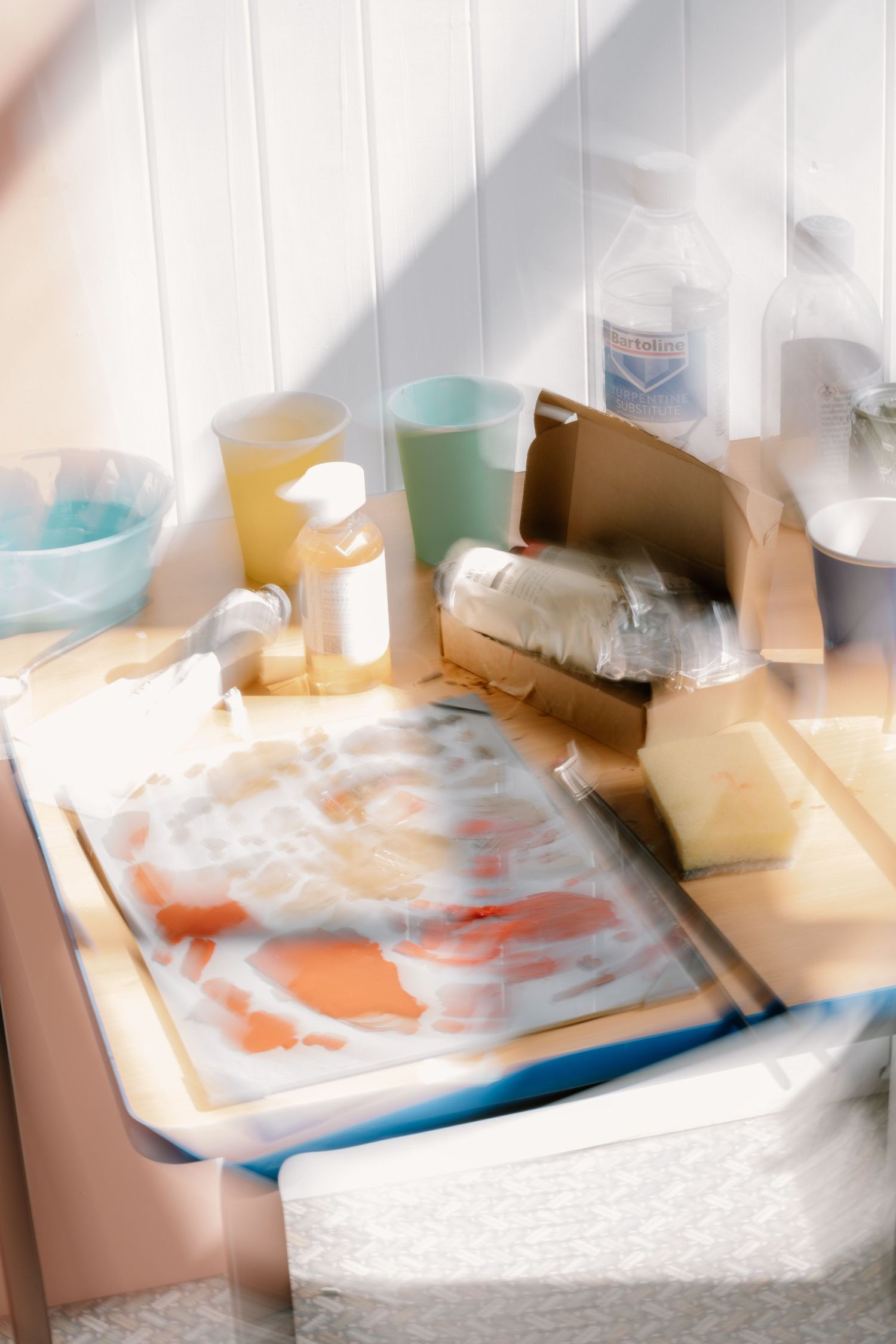 Cluttered craft table with soap making supplies, cardboard box, plastic wrap, and colorful liquids in bright natural light.