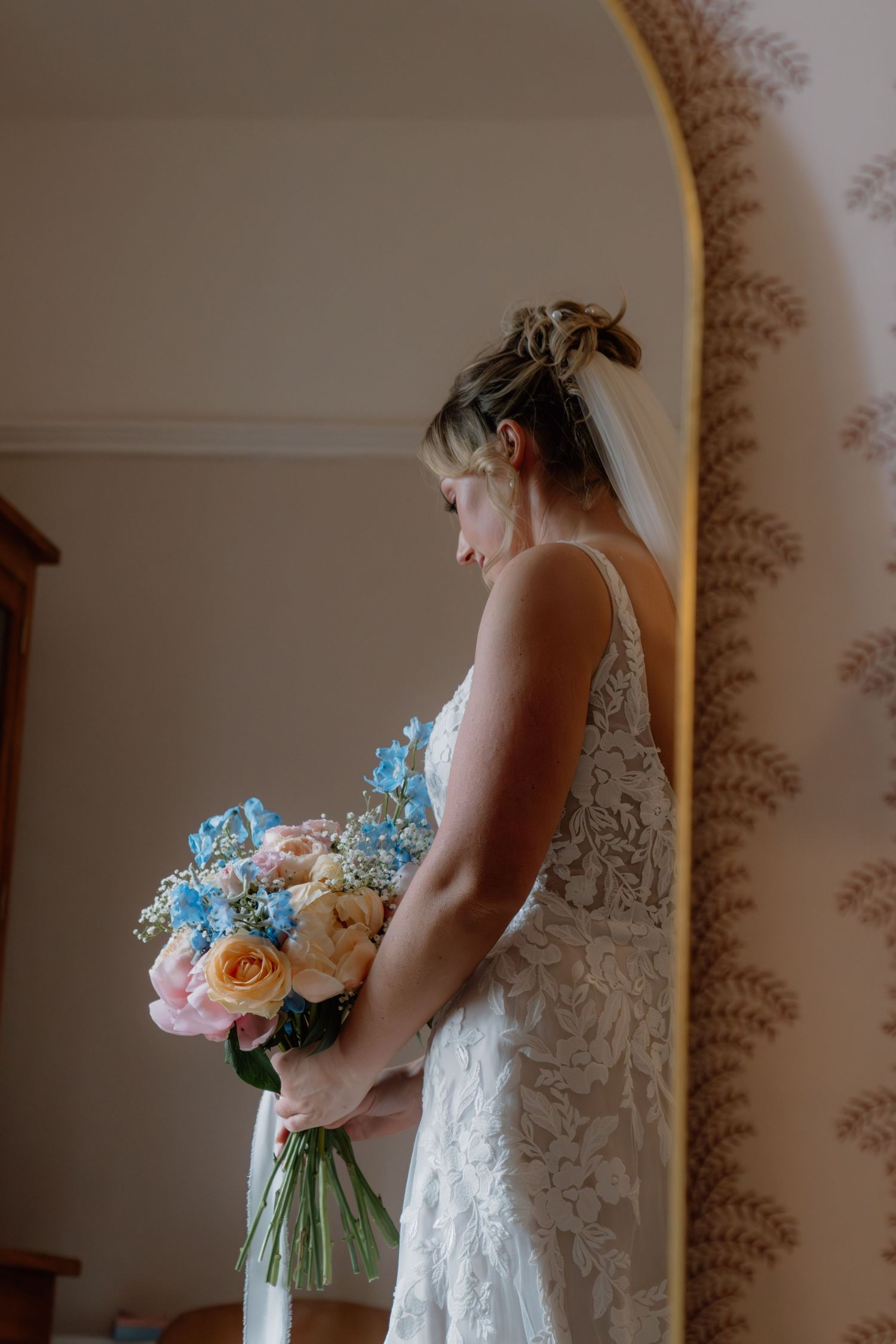 Bride in lace wedding dress holding blue and pink floral bouquet, standing by mirror with veil and updo hairstyle.