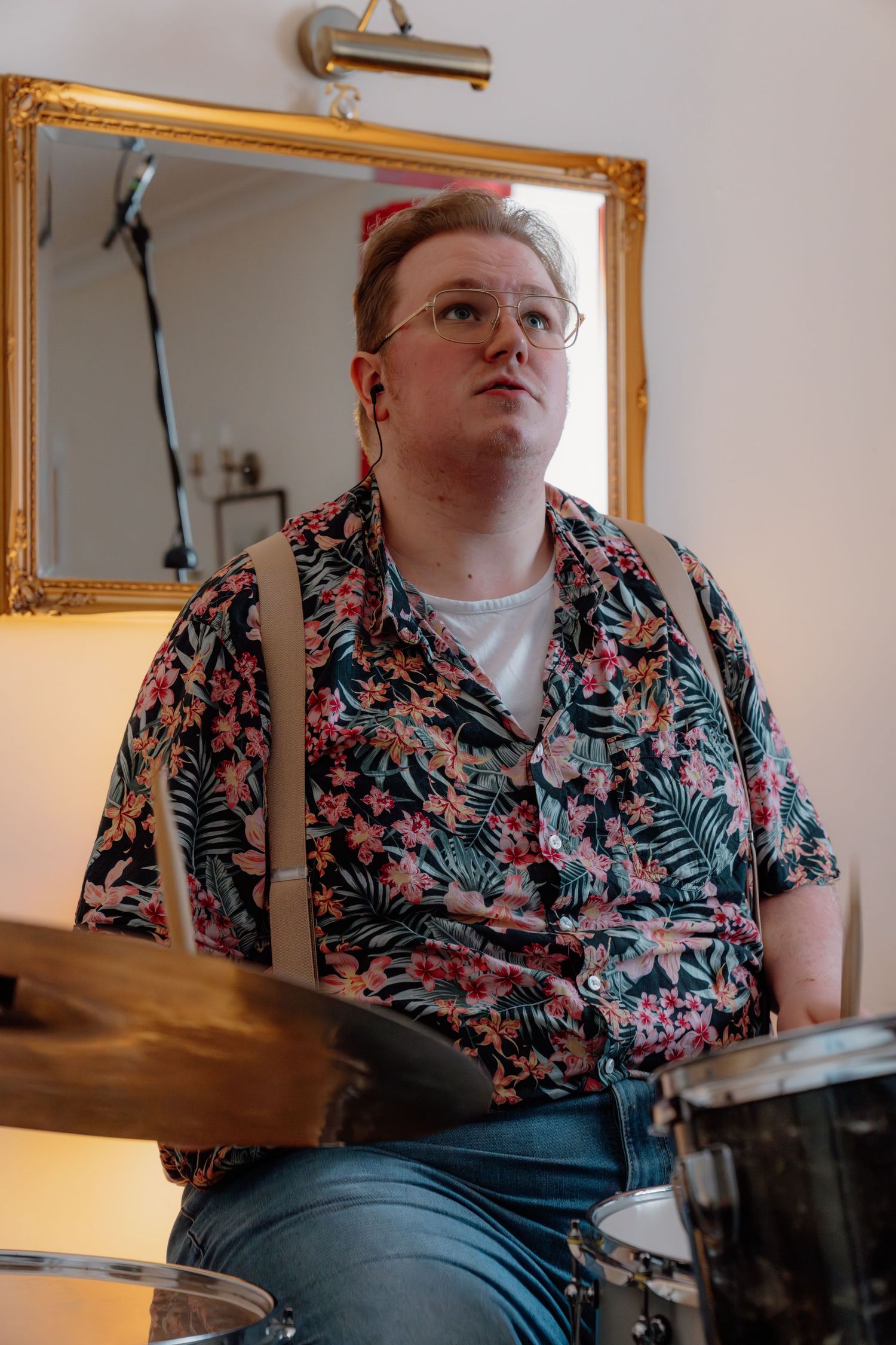 Person in floral shirt playing drums indoors, seated before a gold-framed mirror.