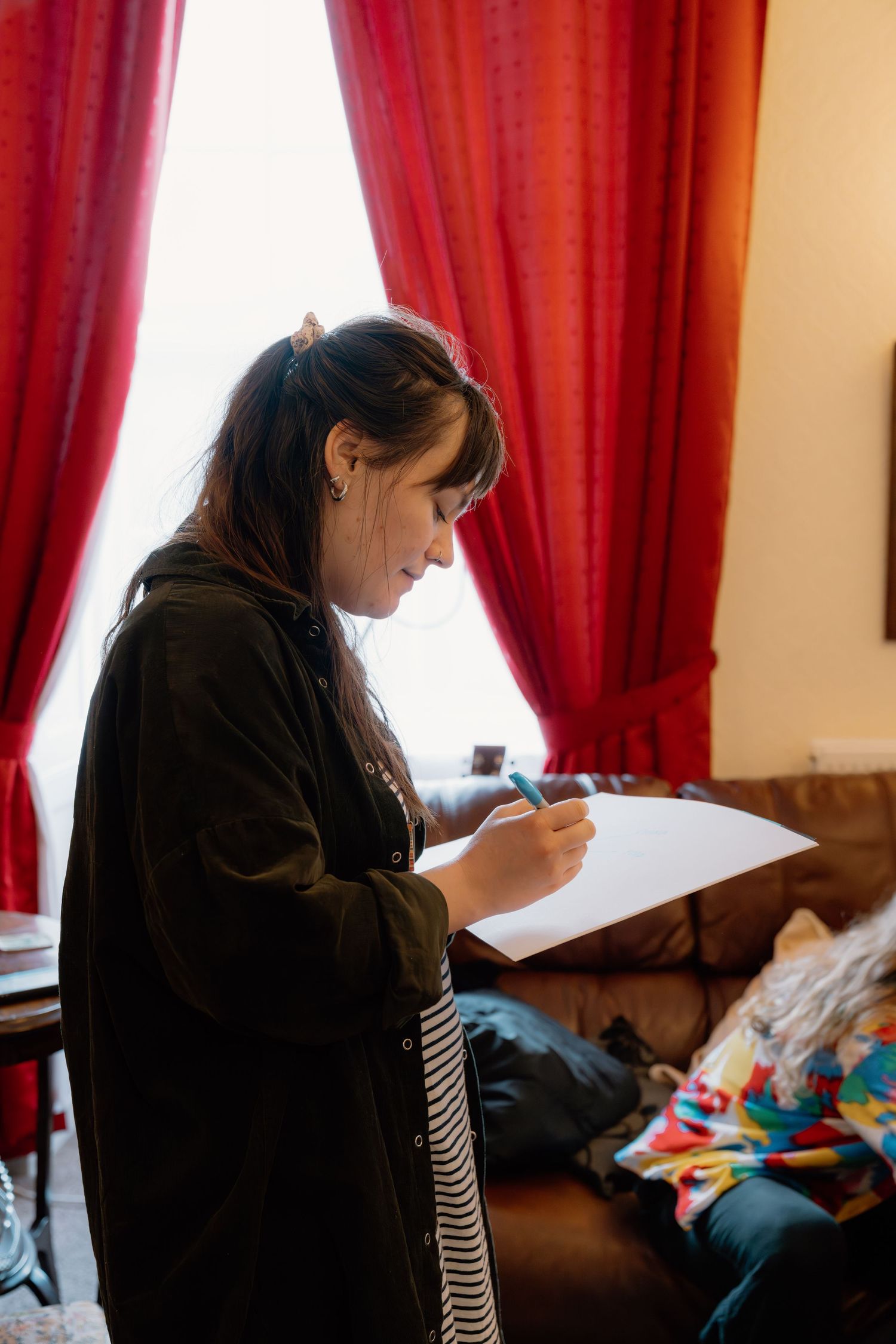 Young woman with dark hair writing on paper with a blue pen, standing near red curtains in a cozy room.