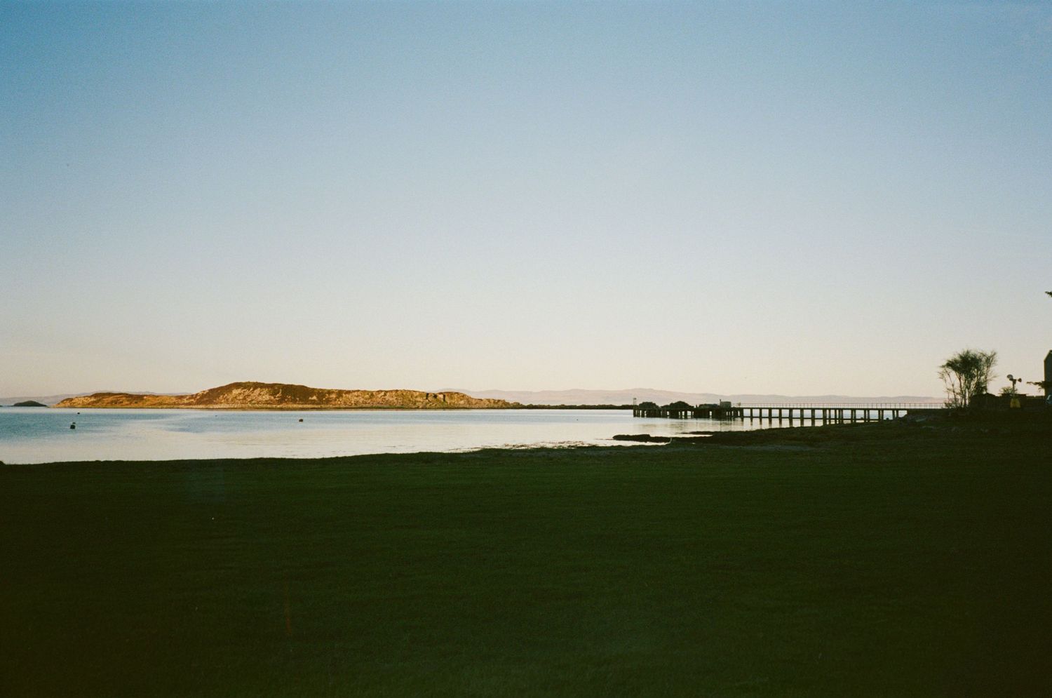 Scenic coastal landscape with calm water, distant hills, and a long pier under a clear blue sky.