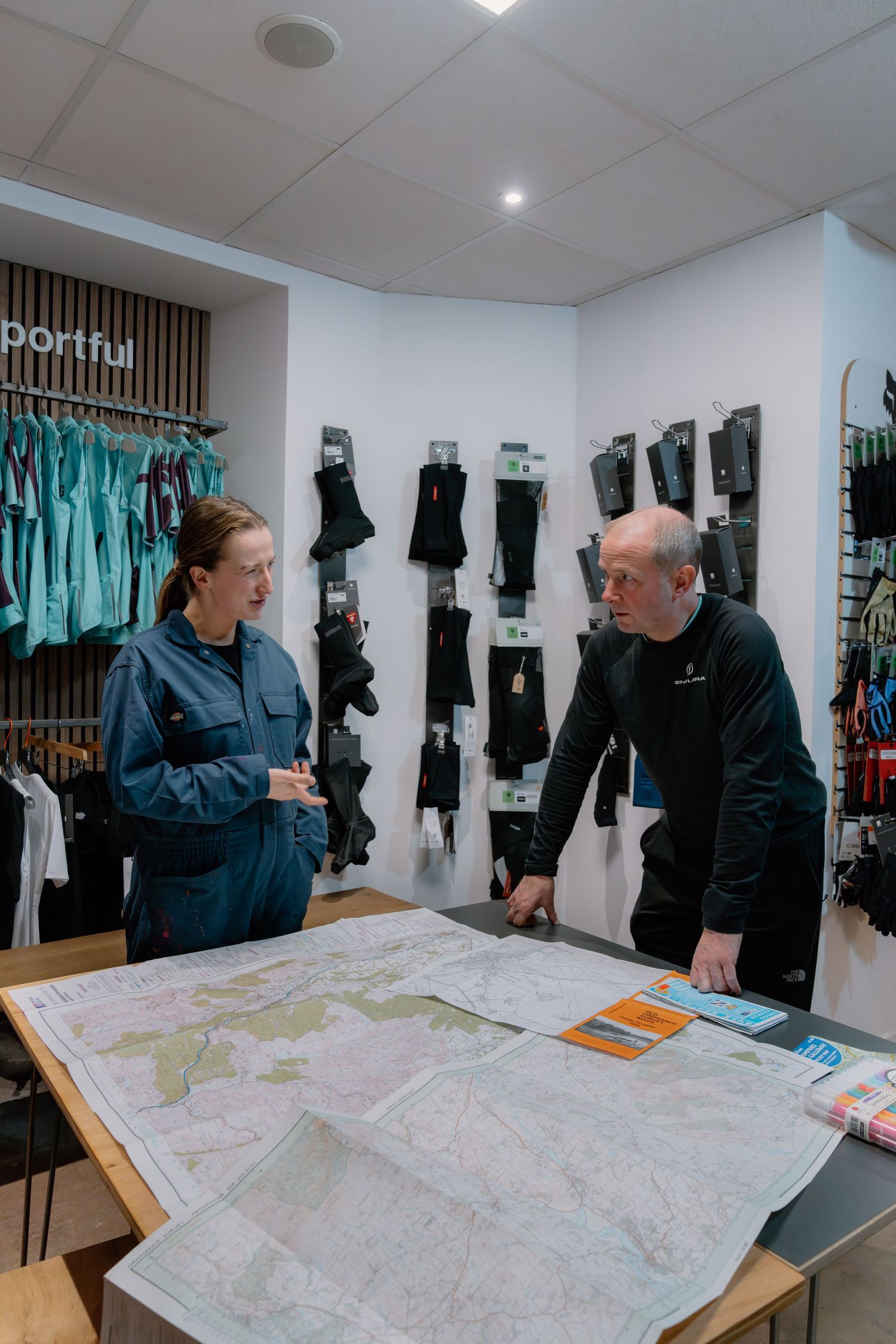 Two people examining a large map on a table inside a sporting goods store with cycling apparel on display.