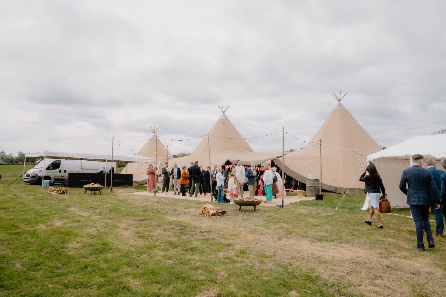 Guests gather outside large tipi tents on a green field at an outdoor wedding venue under a cloudy sky.