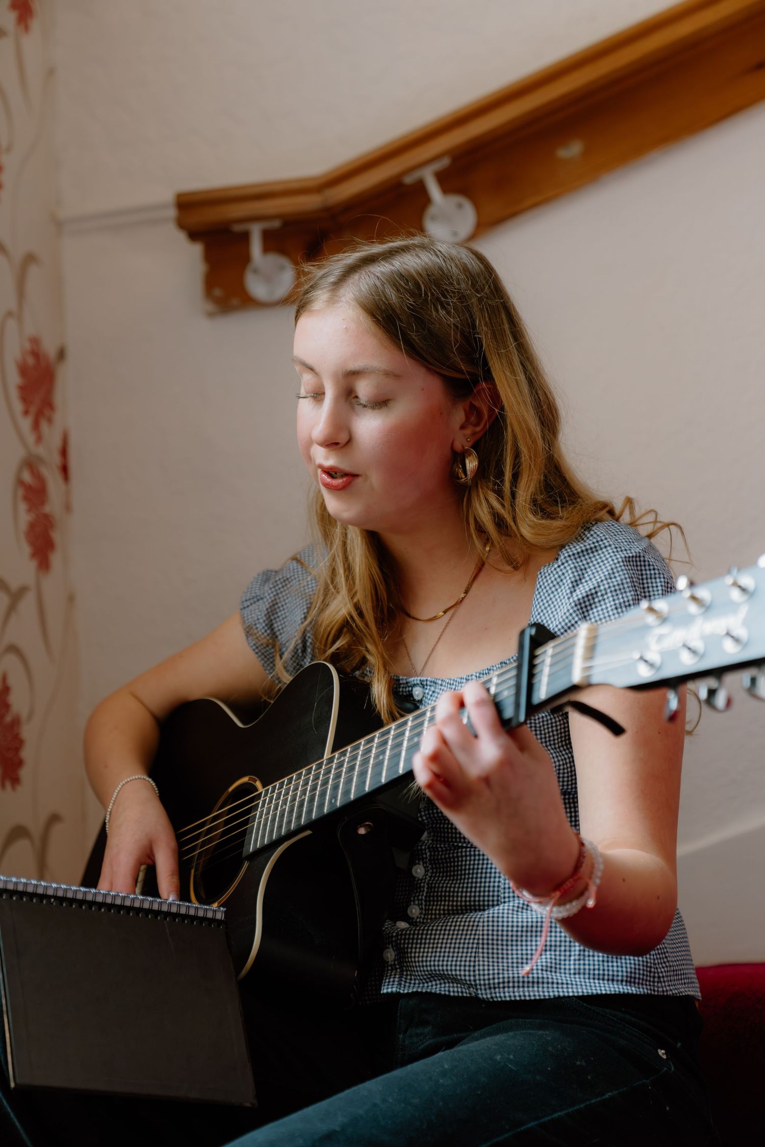 Young girl playing acoustic guitar while singing, seated indoors with sheet music, wearing a blue gingham top.