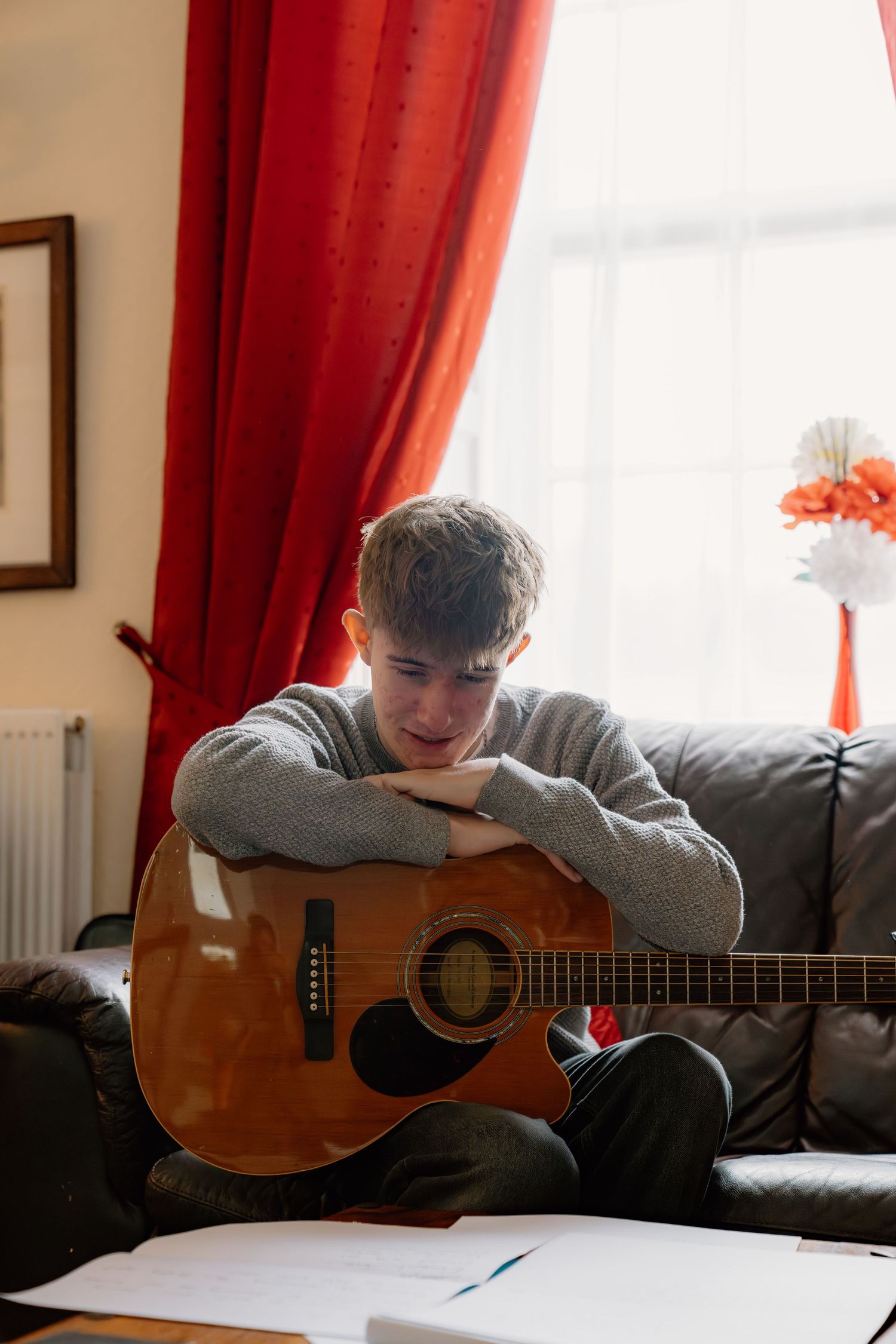 A teenage boy rests his chin on an acoustic guitar while sitting on a leather sofa near red curtains.