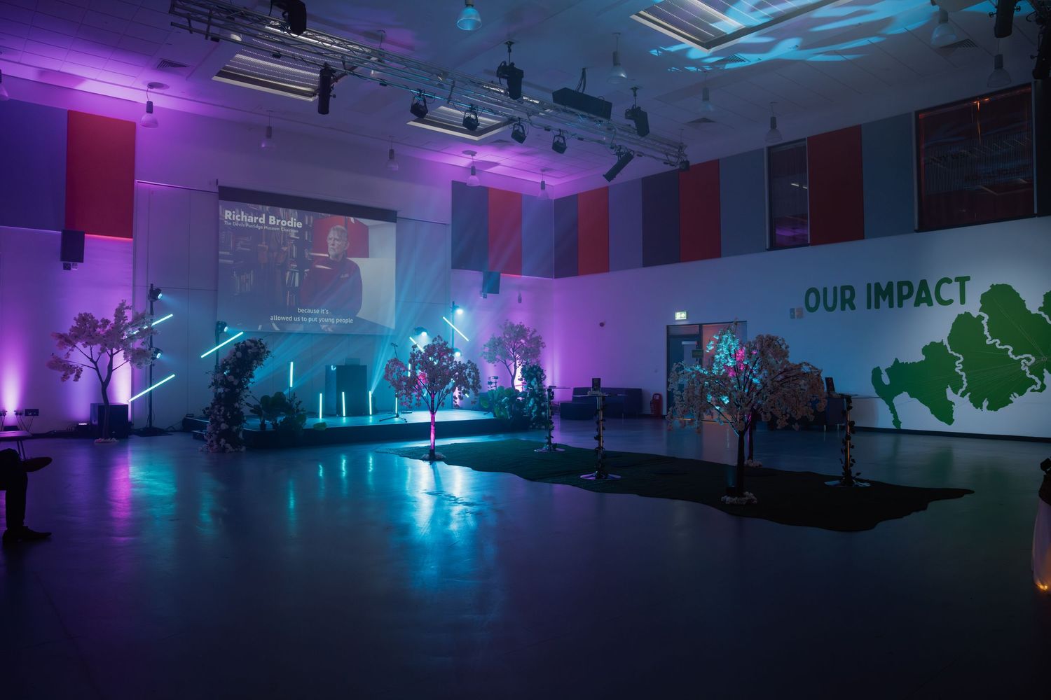 A dramatic event space with purple and teal lighting, decorated trees, a projection screen, and an 'Our Impact' green mural on the wall.