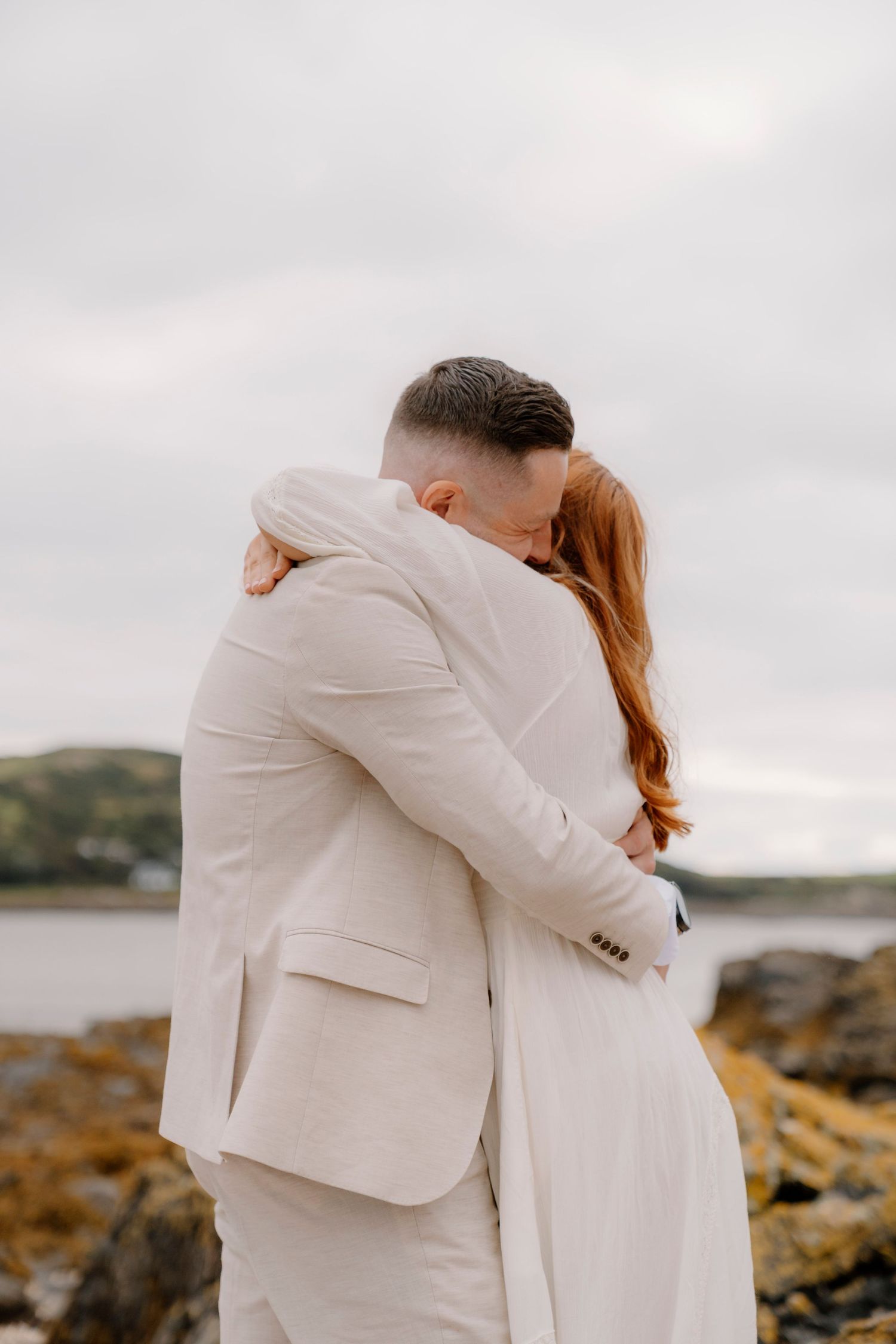 Couple in white formal wear embrace on rocky coastal overlook.