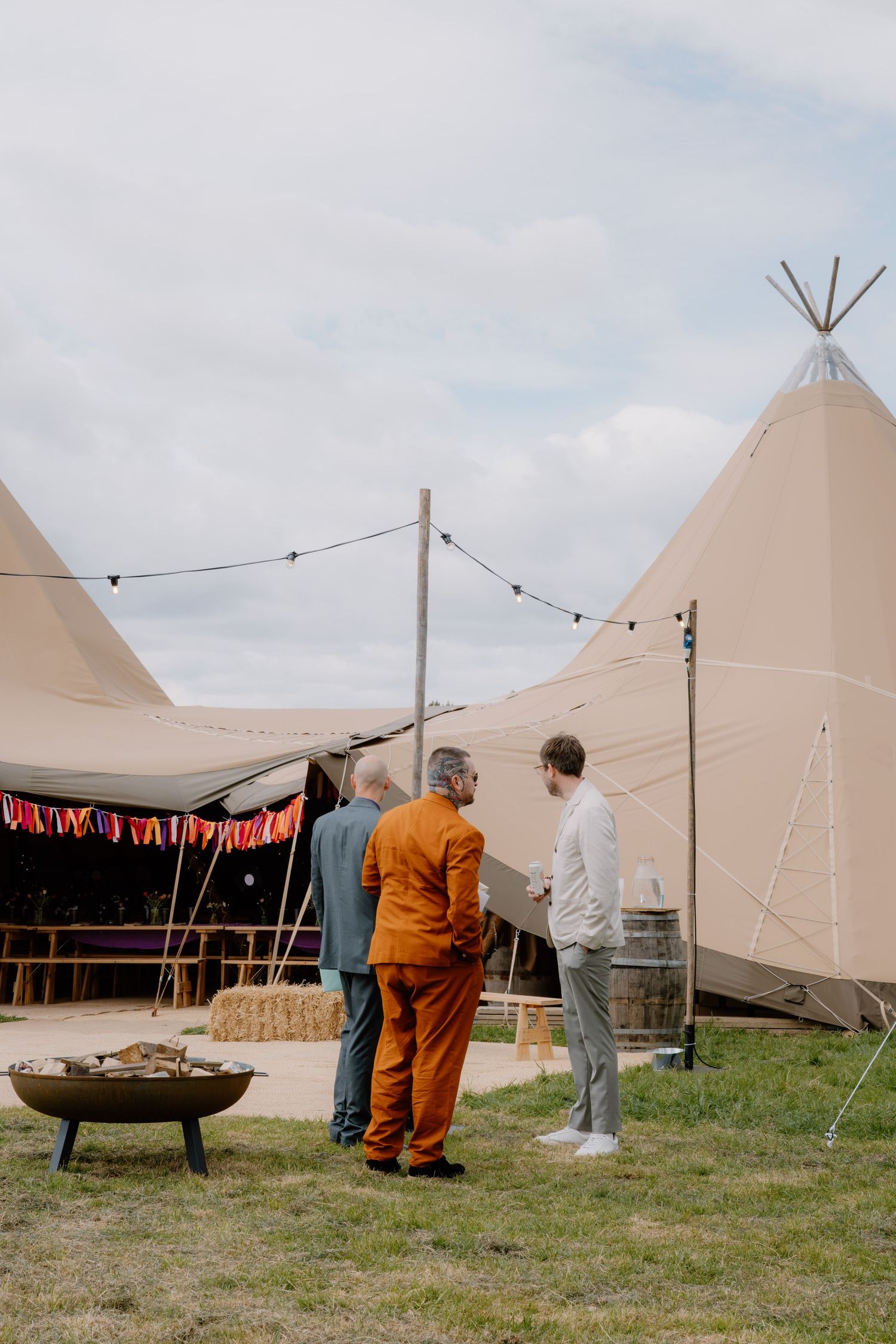 Three men conversing outside large tipi tents at an outdoor festival event with string lights and a fire pit.