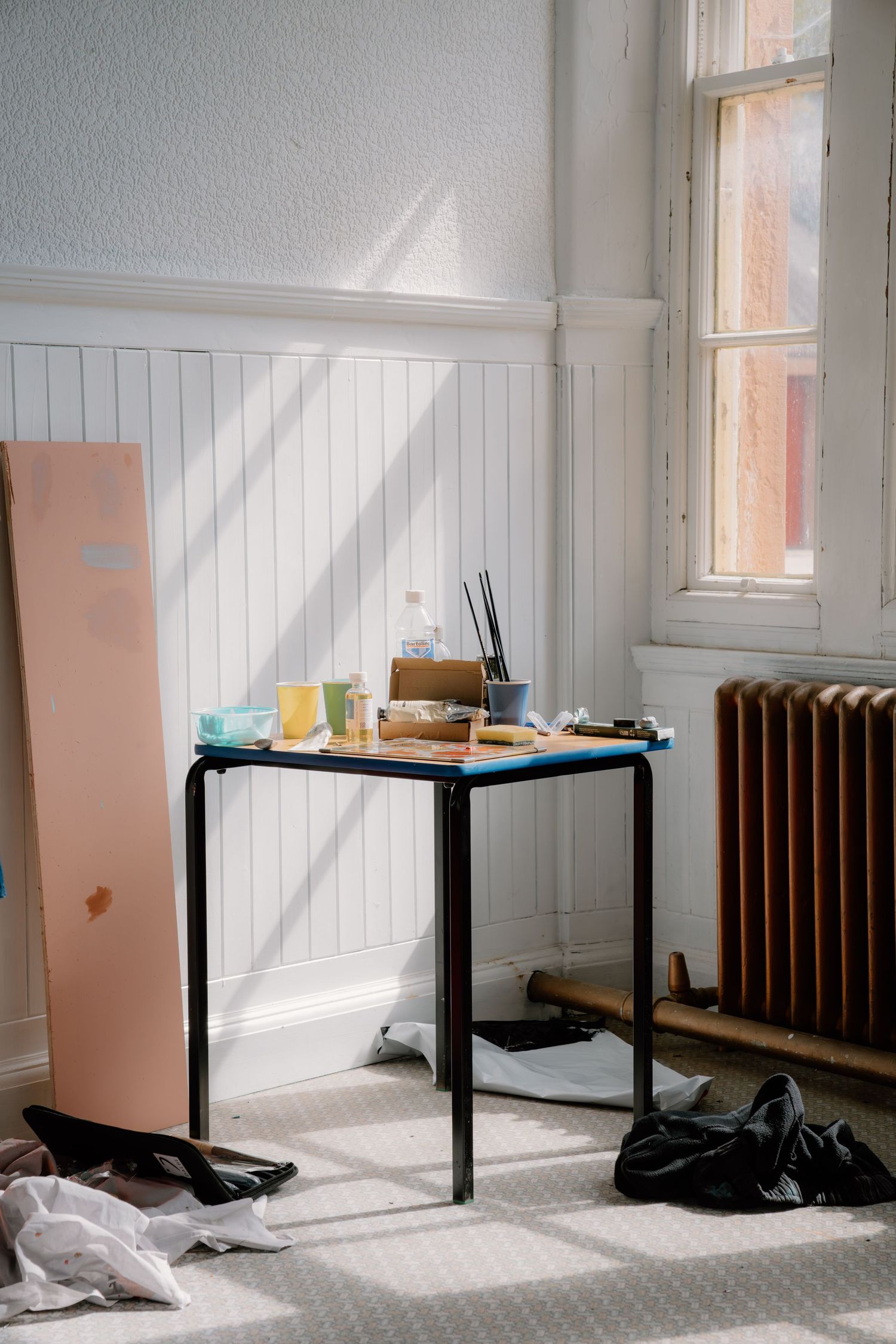 Cluttered artist studio desk with paint brushes and supplies near a window with a cast iron radiator.