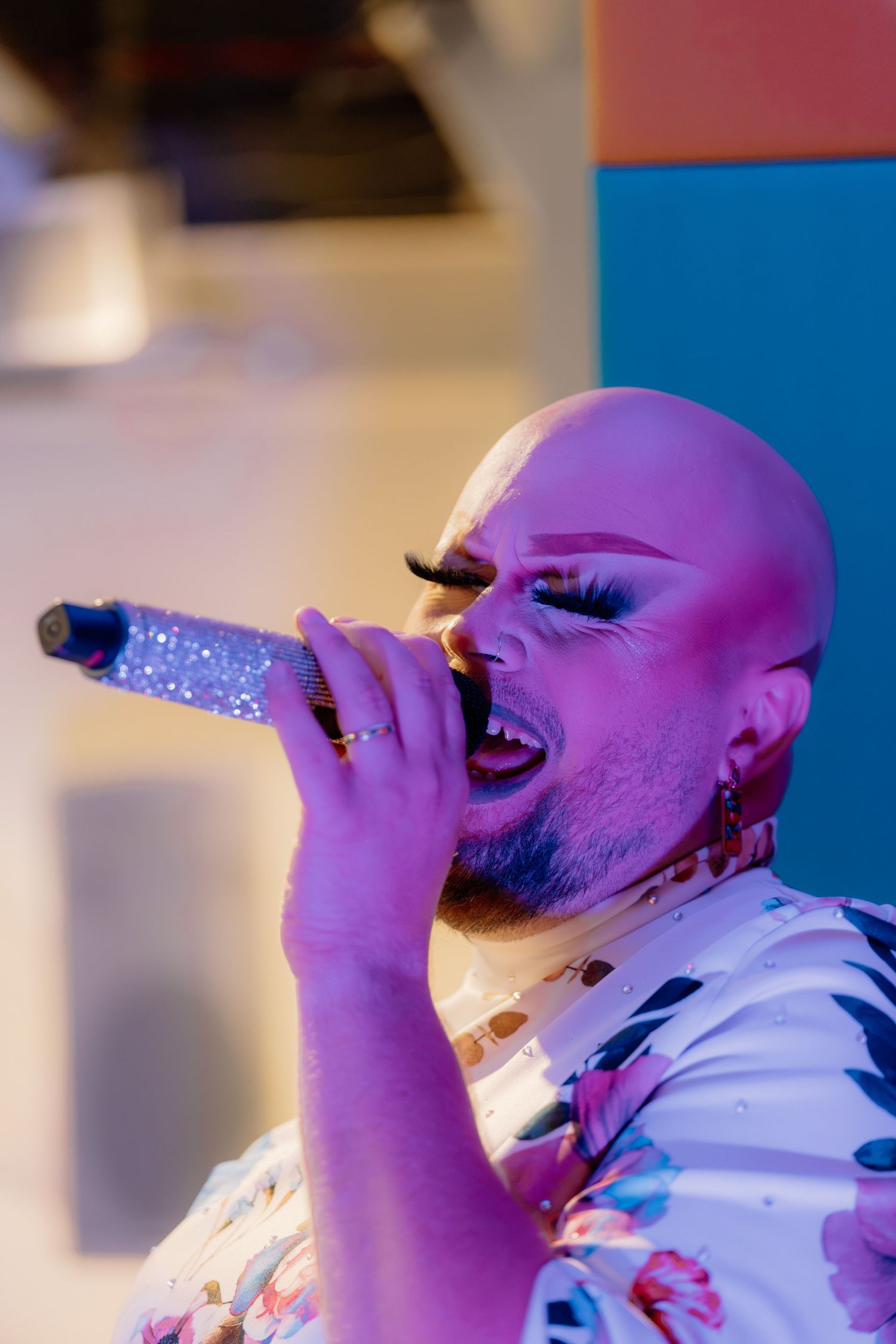 Bald drag performer with dramatic makeup singing passionately into a glittery microphone under purple stage lighting.