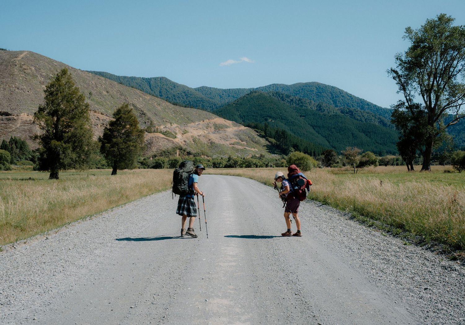 Hikers with backpacks traverse a gravel mountain road on a sunny day with rolling hills in the background.
