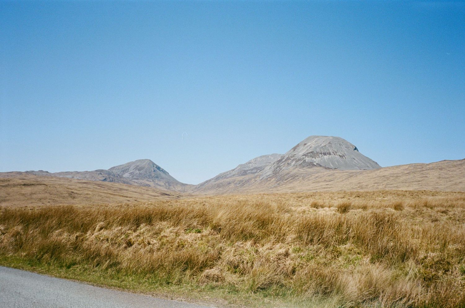 Scenic highland landscape with golden grasslands, a winding road, and two rugged mountain peaks under a clear blue sky.