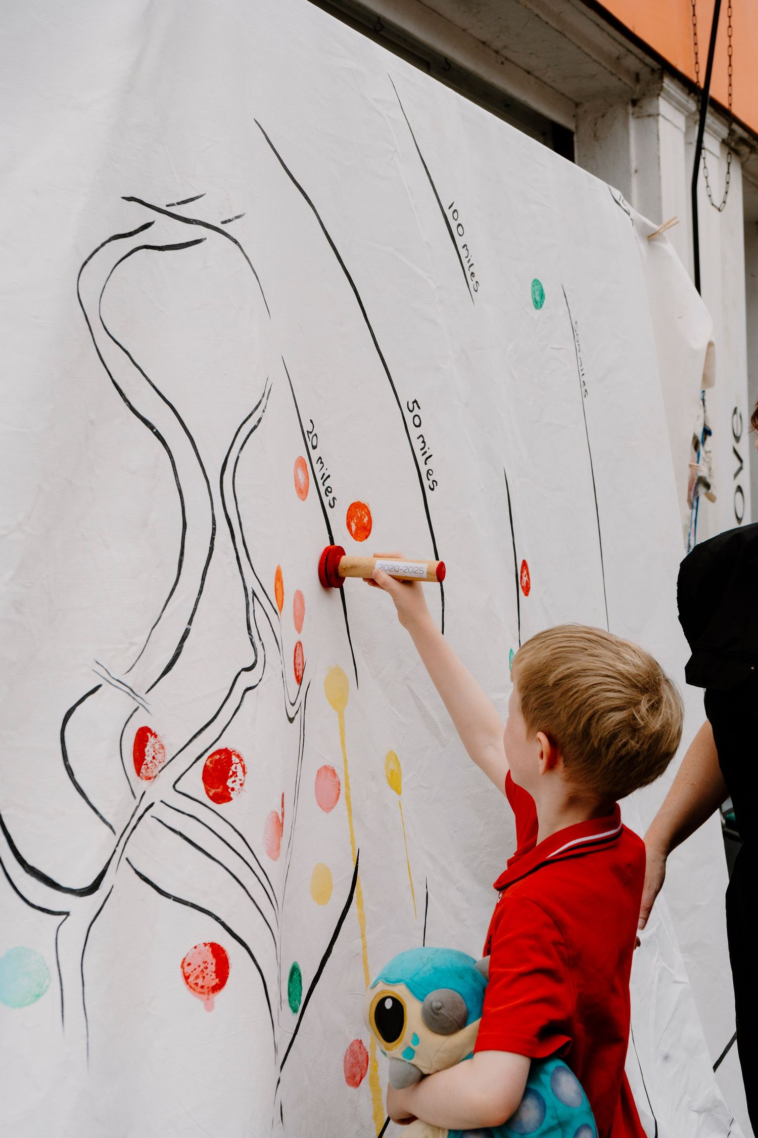 Young boy in red shirt stamping colorful dots on a large white mural wall while holding a plush toy.