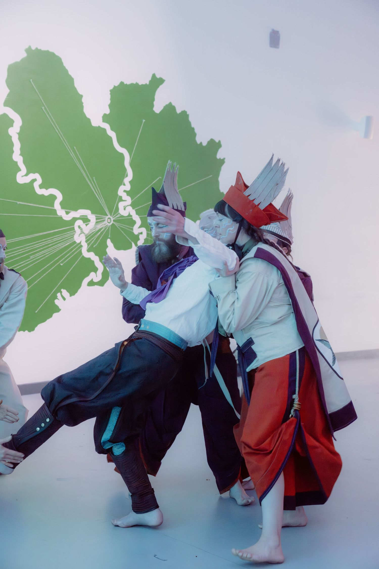 Two cosplayers in colorful fantasy costumes perform a dramatic scene in front of a green map mural.