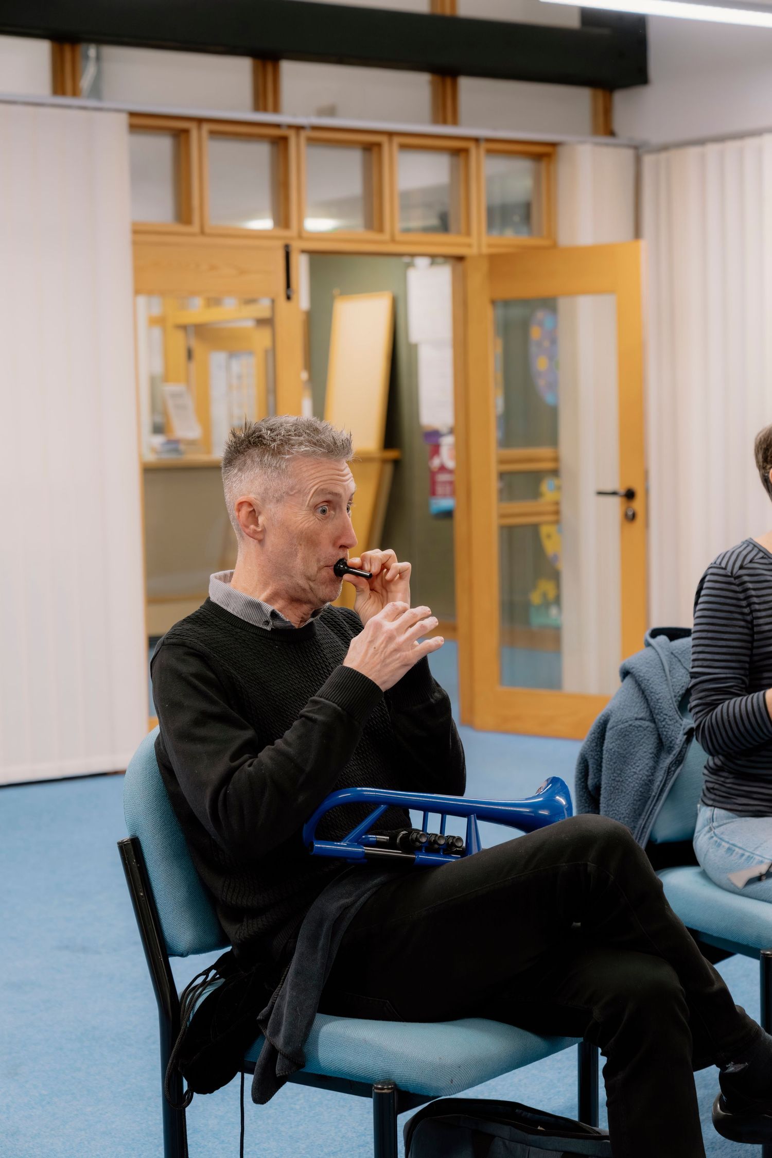 Middle-aged man in black sweater playing a small instrument while seated, with a blue trumpet resting on his lap indoors.