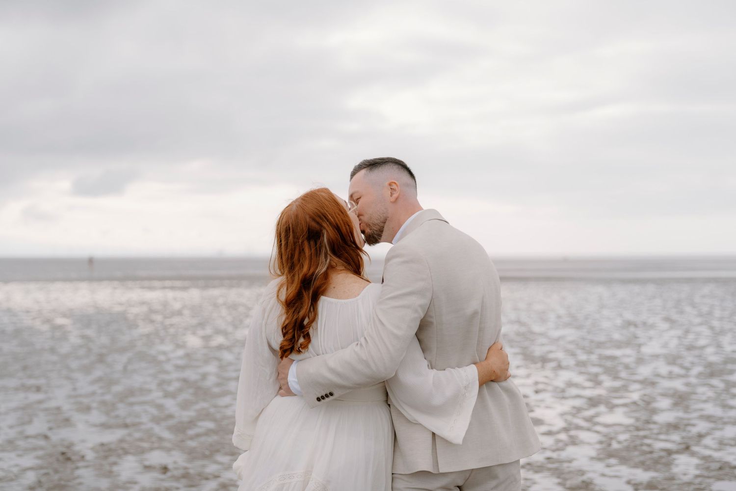 Romantic couple embraces on a misty beach during an overcast day.