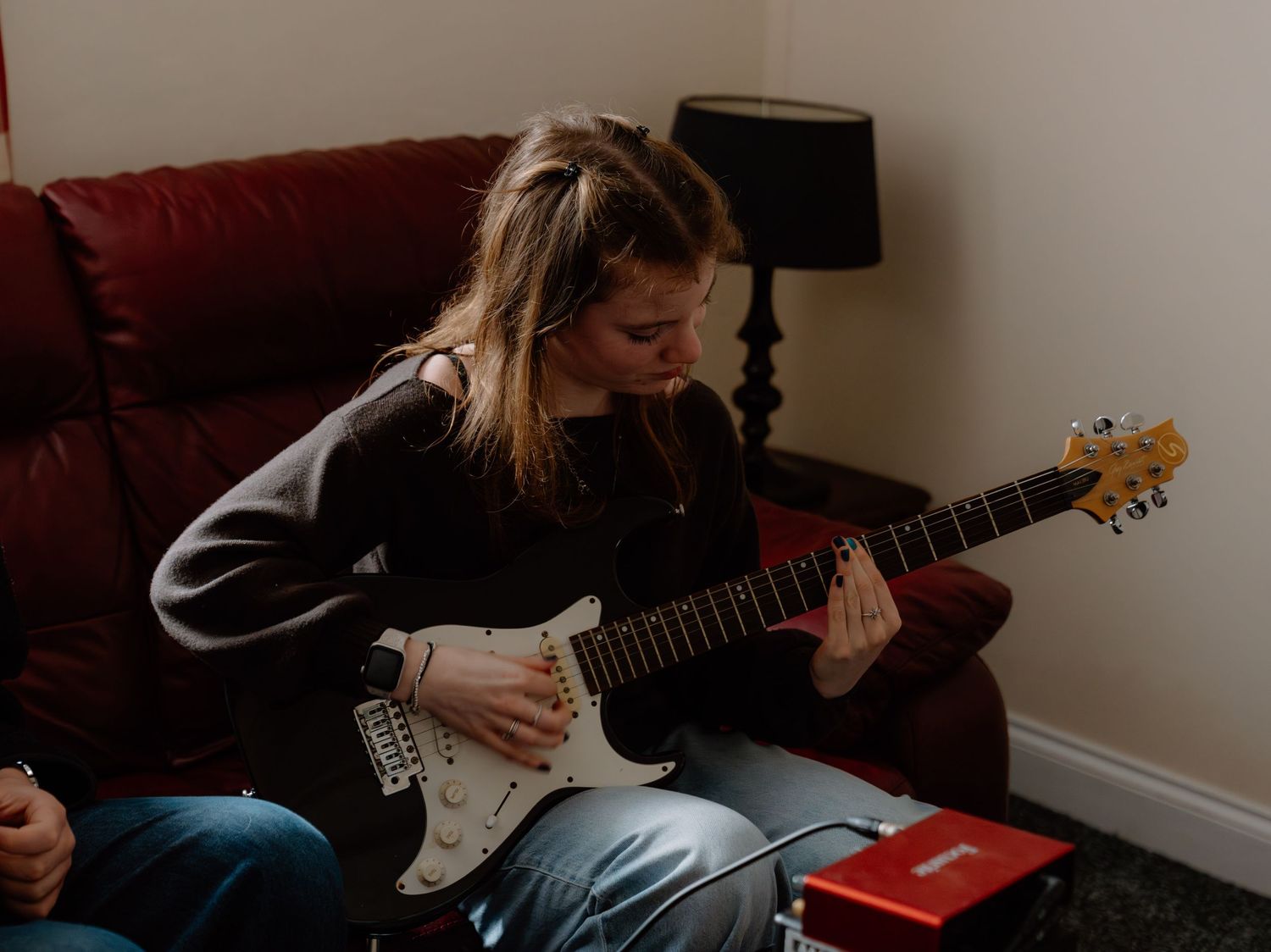 Young woman playing electric guitar while sitting on red sofa in cozy living room.