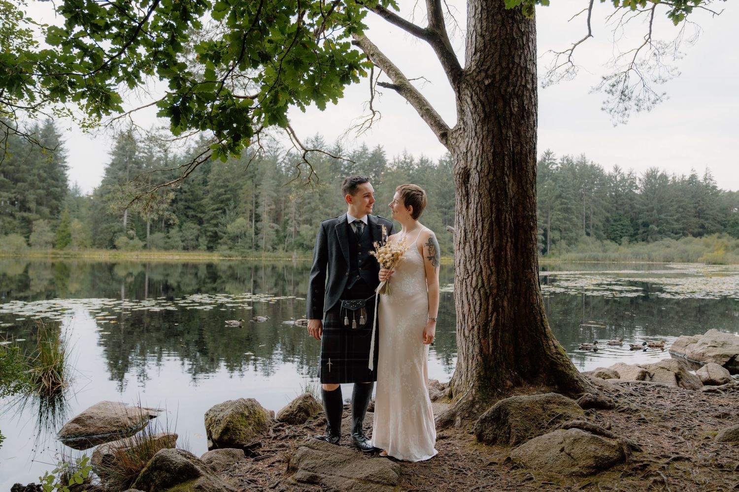 Couple in Scottish wedding attire stands by a tranquil loch surrounded by pine trees, groom in kilt, bride holding bouquet.