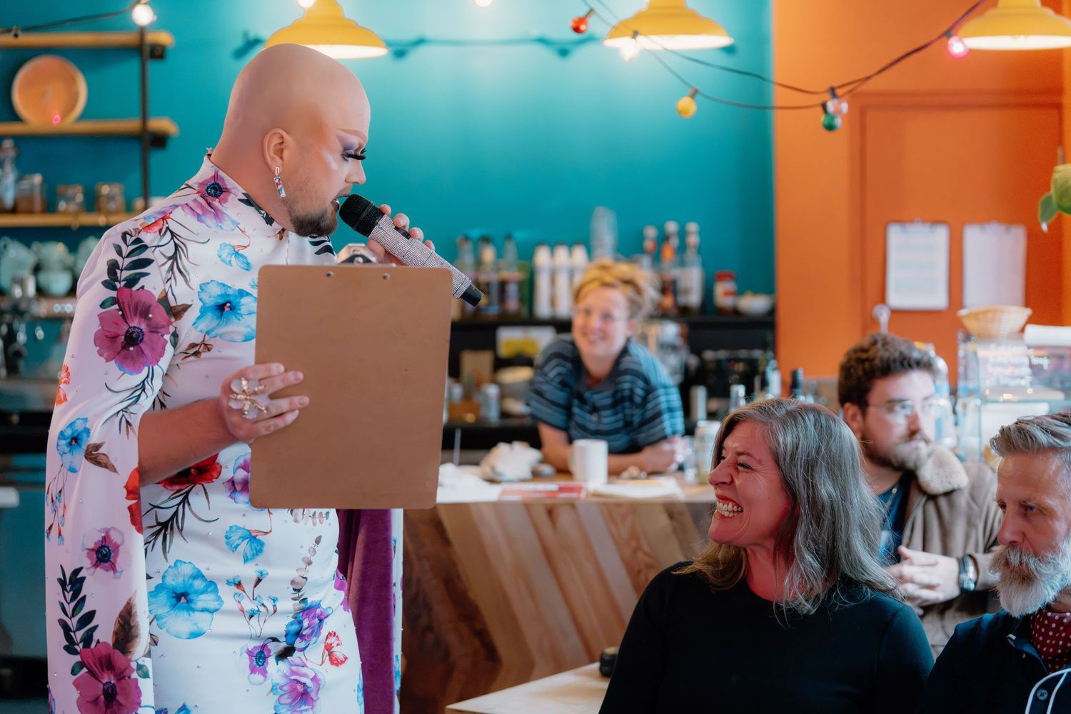 Bald woman in floral dress presents award to smiling guest in colorful cafe setting.