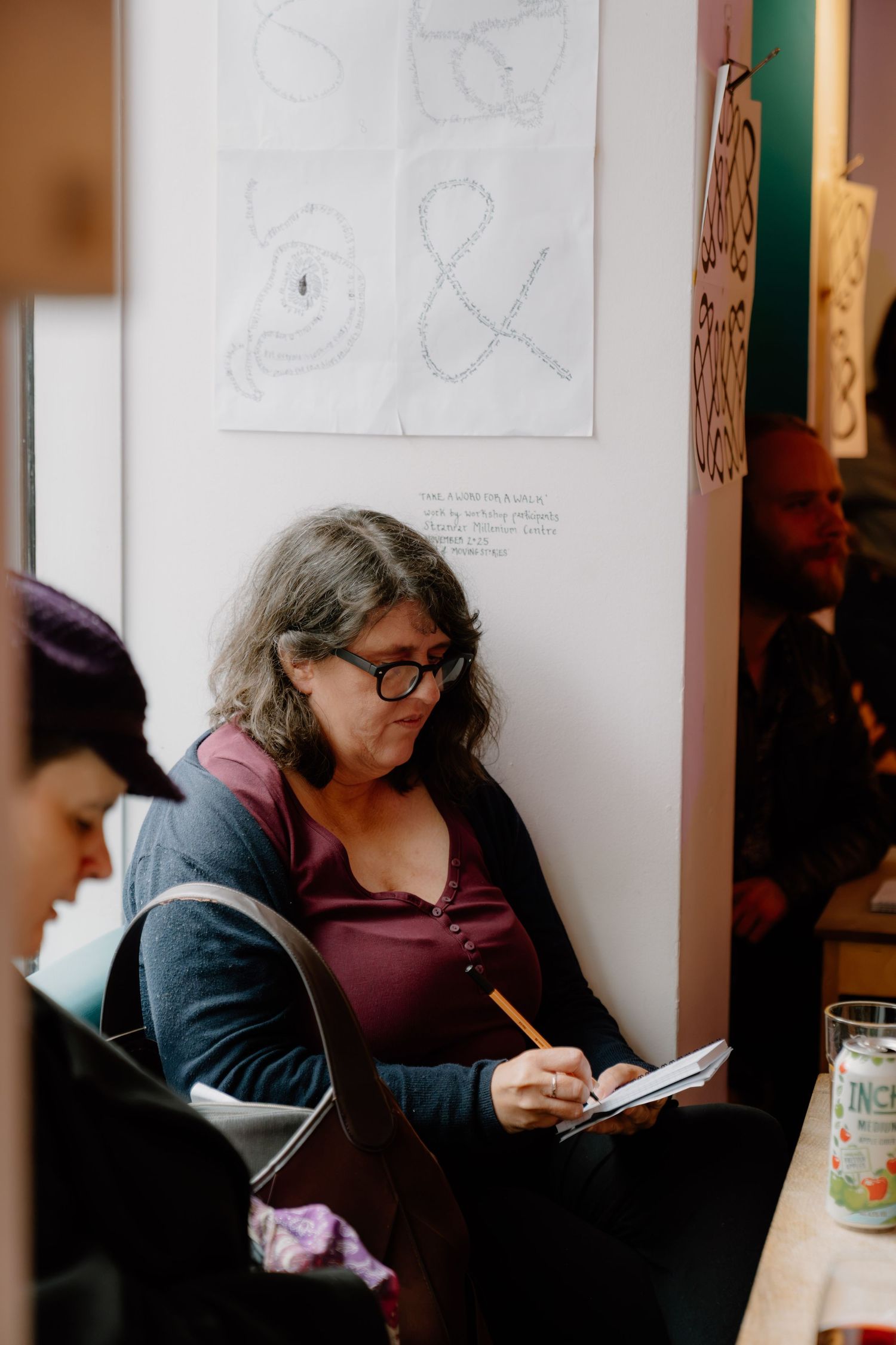 Woman with glasses writing in notebook at cafe, sketches on wall behind her.