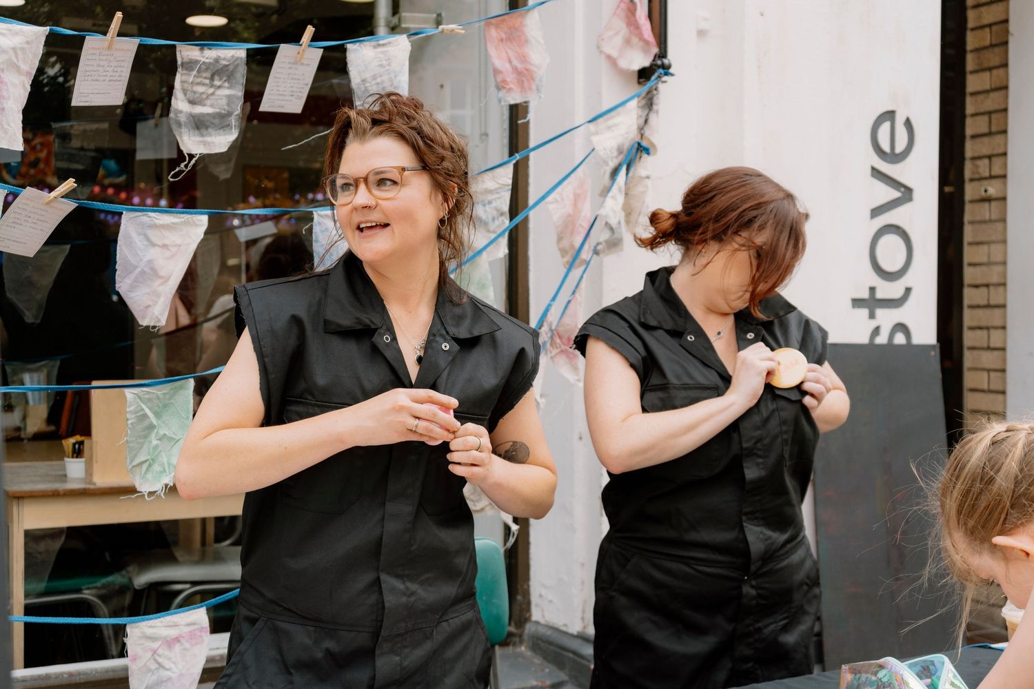 Two women in black sleeveless shirts stand outdoors near a shop with bunting decorations overhead.