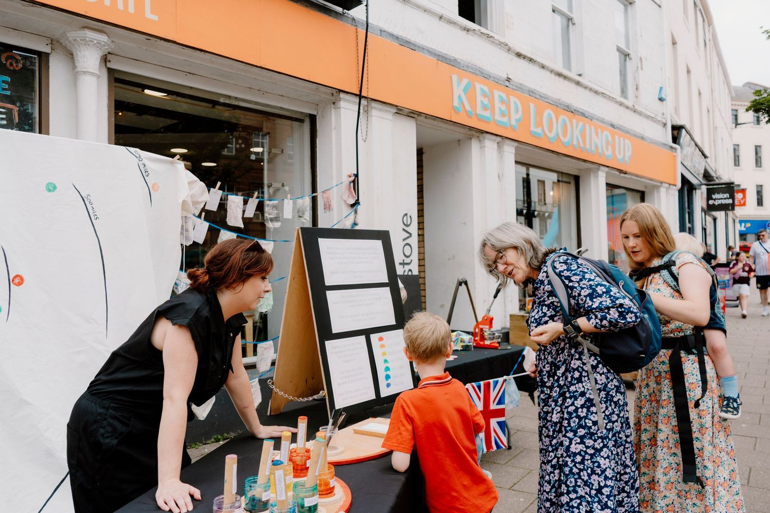 People browsing outdoor market stalls on a sunny street outside a shop with a Keep Looking sign.