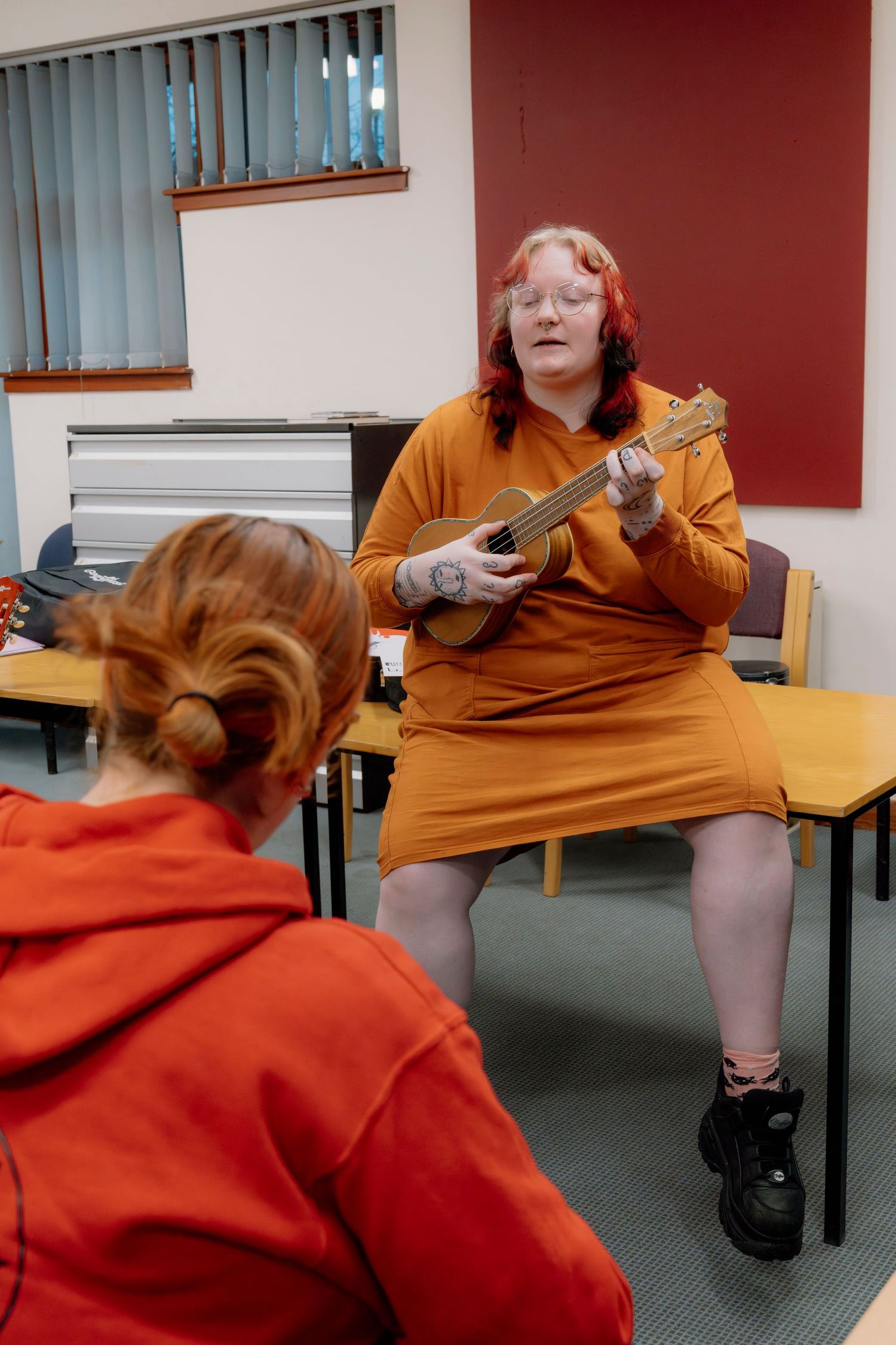 Woman in orange dress playing ukulele while seated, performing for a student in a classroom music lesson.