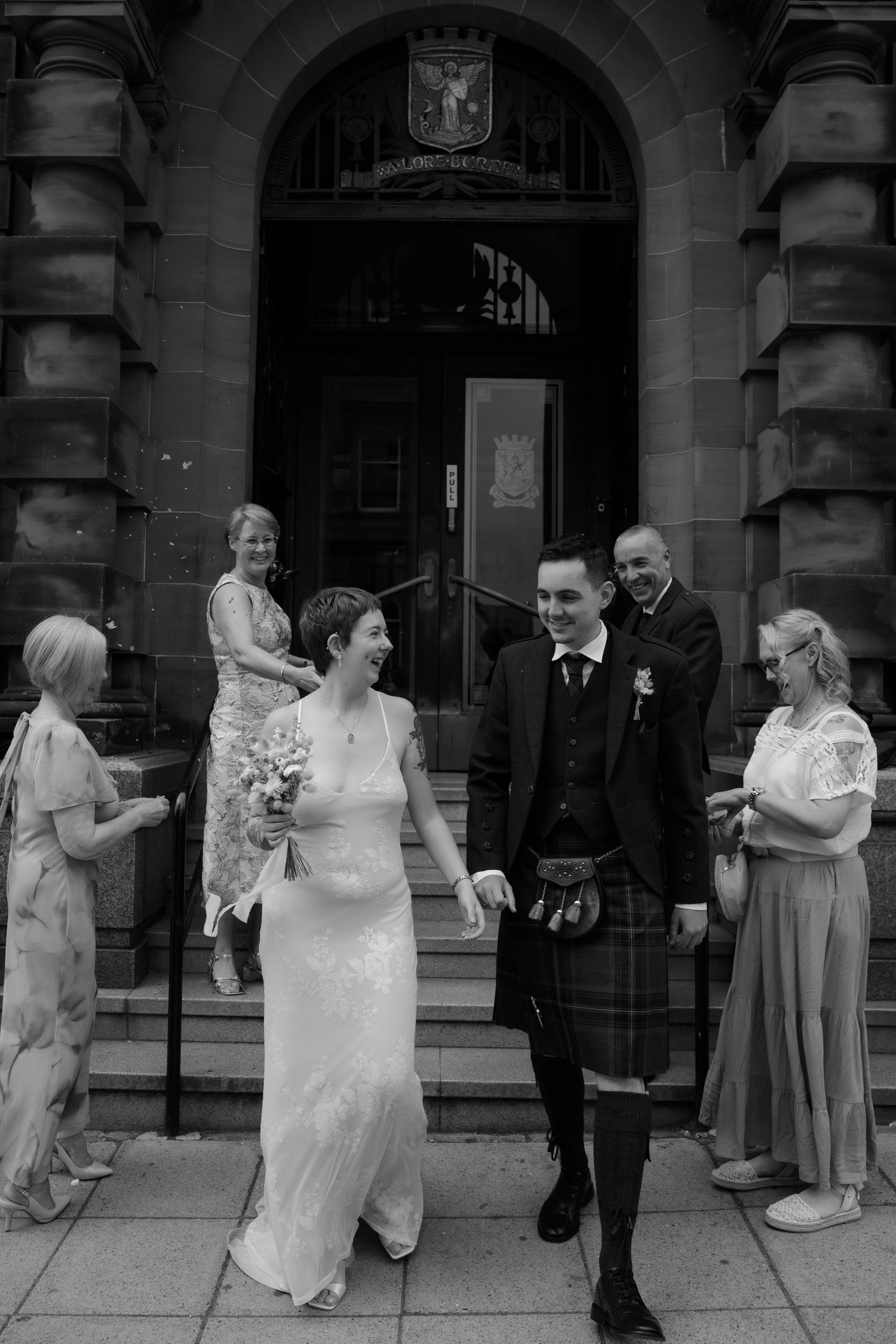 Black and white photo of a smiling bride and groom in kilt leaving a historic building after their wedding ceremony.