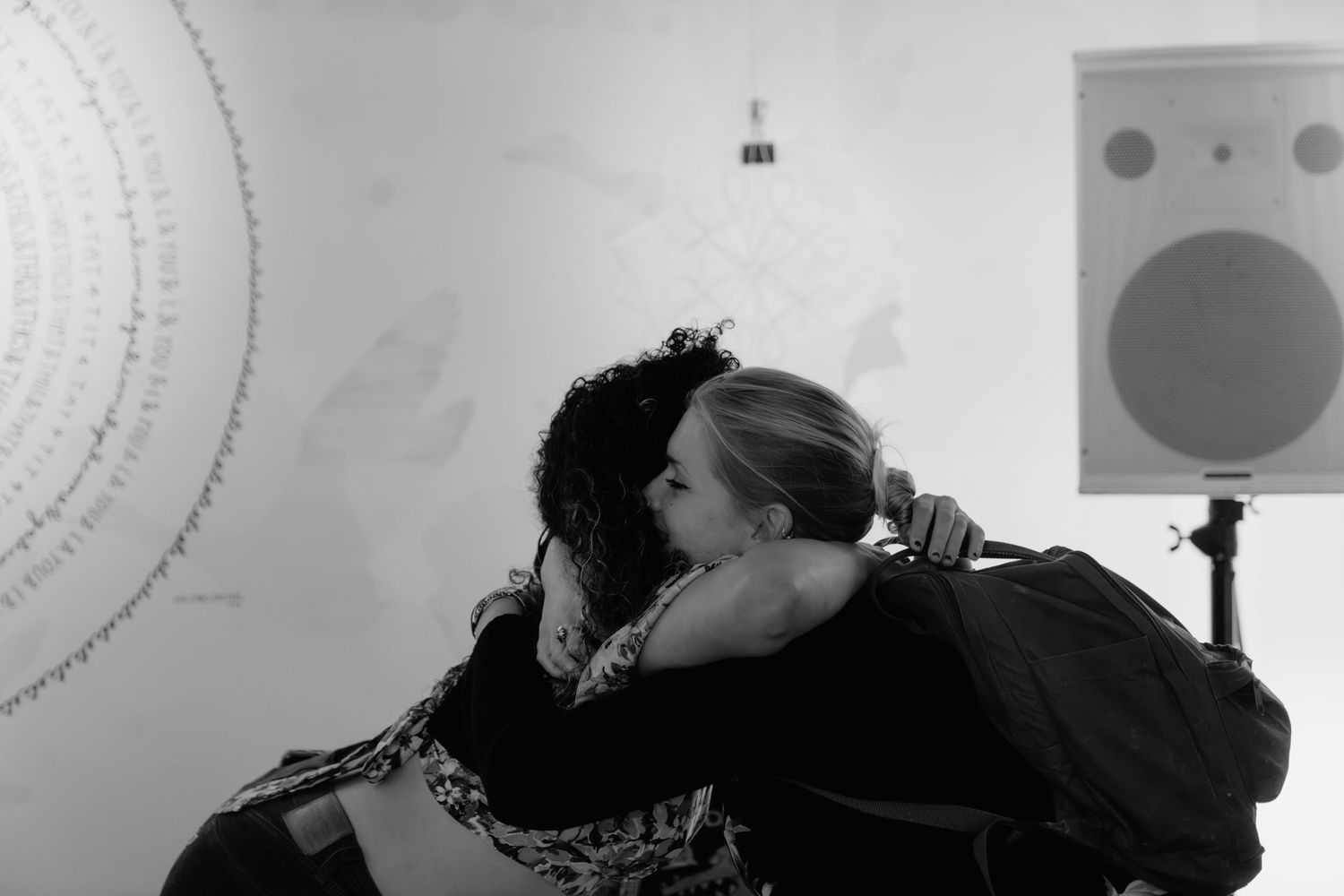 Two women sharing a warm embrace in a black and white photo, with a speaker and wall art visible in the background.