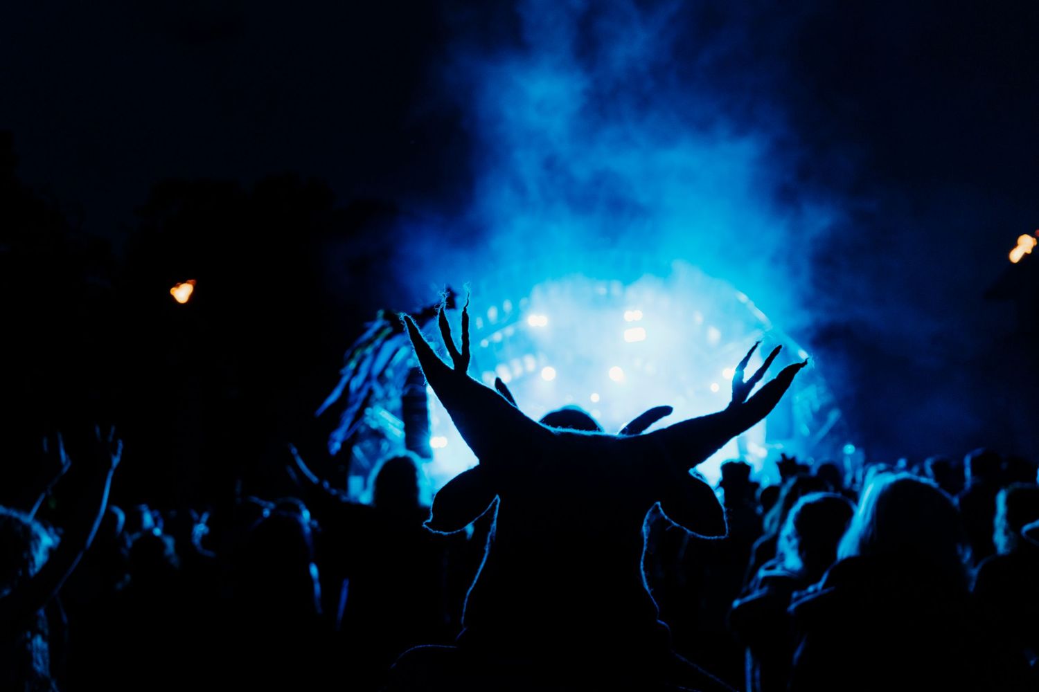 Silhouetted crowd with raised arms at a concert venue illuminated by dramatic blue stage lighting and smoke effects.