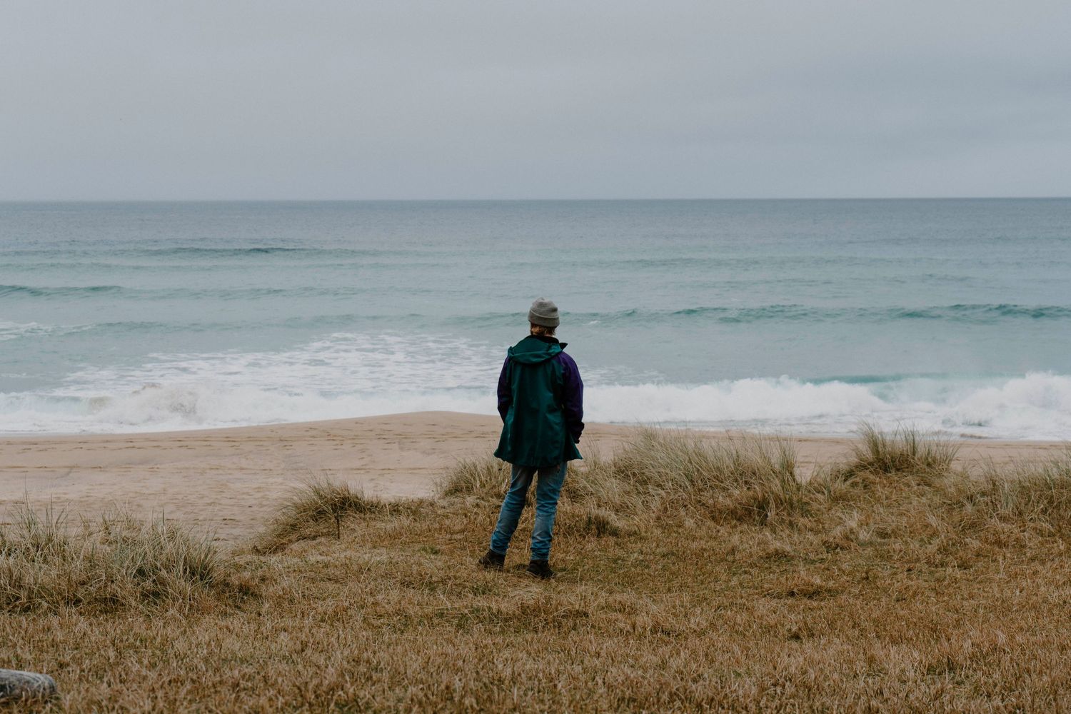 A person stands on a sandy beach overlooking a moody gray ocean on an overcast day.