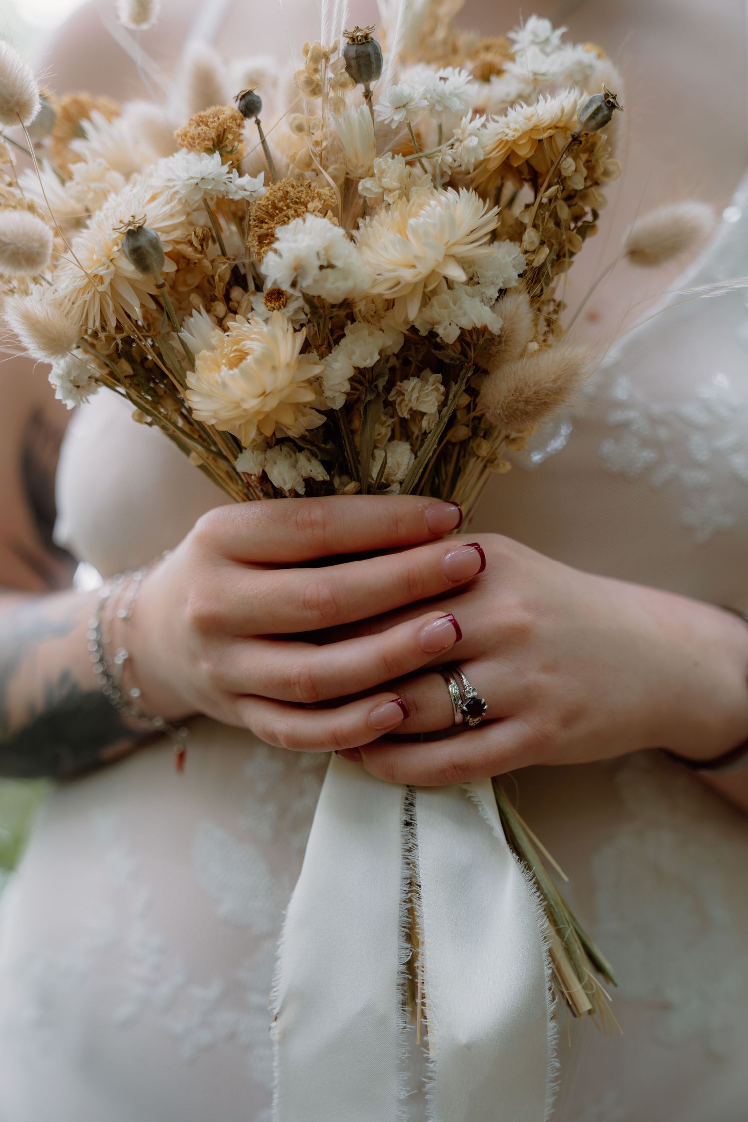 Bride holding a dried floral bouquet with white and tan flowers, wearing a silver ring and bracelet.